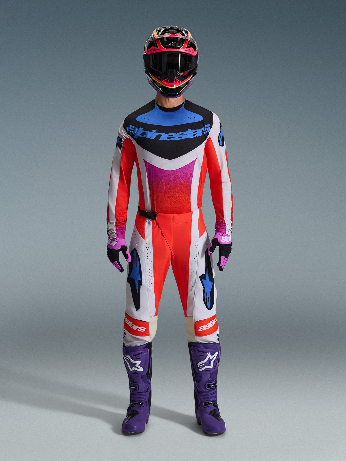 Motorcrossrijder in volledige race-uitrusting, Alpinestars 2026 Techstar Knif Jersey en bijpassende broek in wit en oranje met blauwe en paarse accenten, paarse racelaarzen met wit logo en een meerkleurige helm, gecoördineerde outfit voor off-road competitie