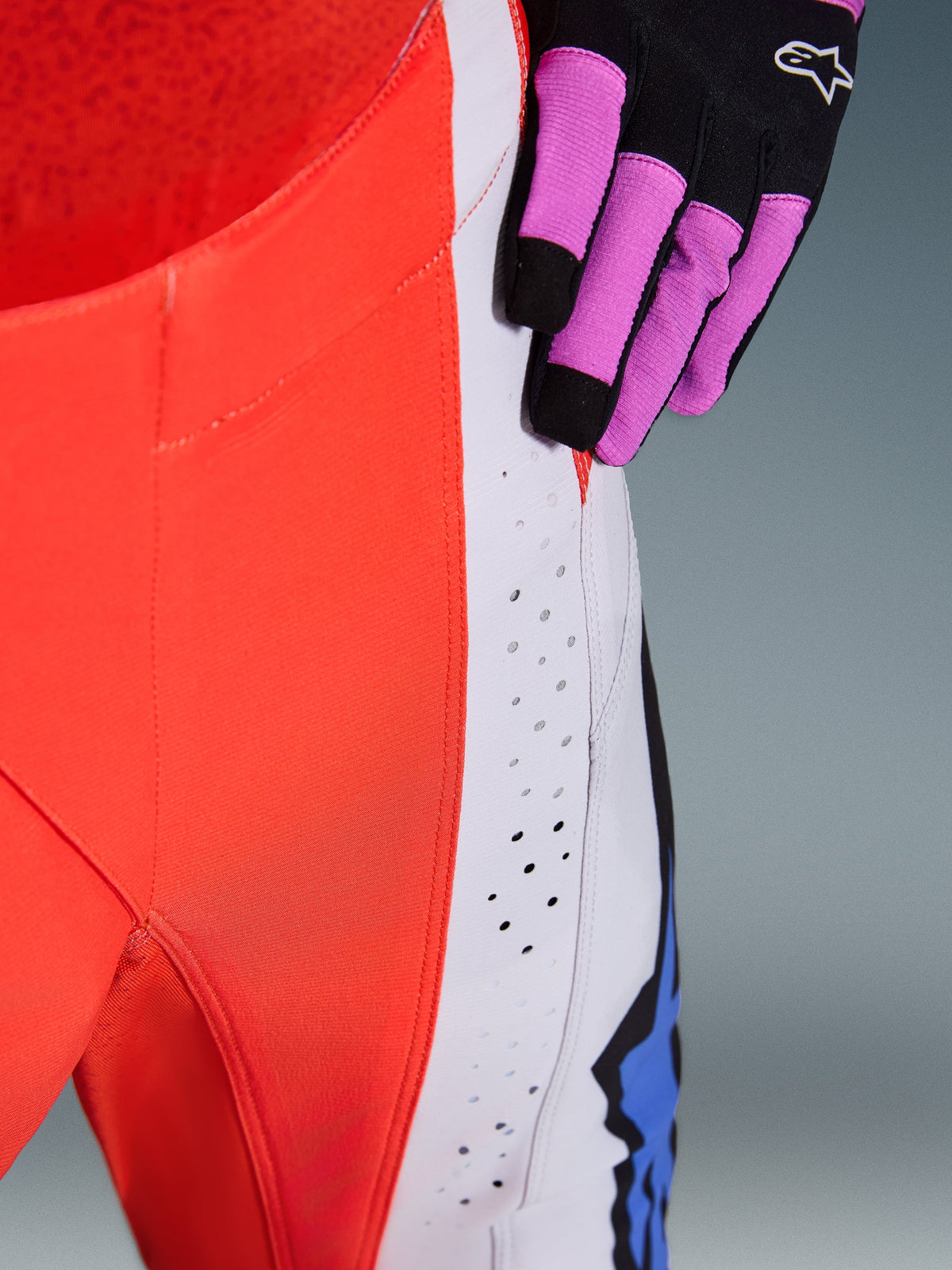 Alpinestars 2026 Techstar Knif broek, close-up van heup en zijpaneel in Orange Fluo en Light Gray, voorzien van lasergesneden perforaties voor luchtstroom, gedragen met een zwart-paarse motorcrosshandschoen, hoogwaardige race-uitrusting