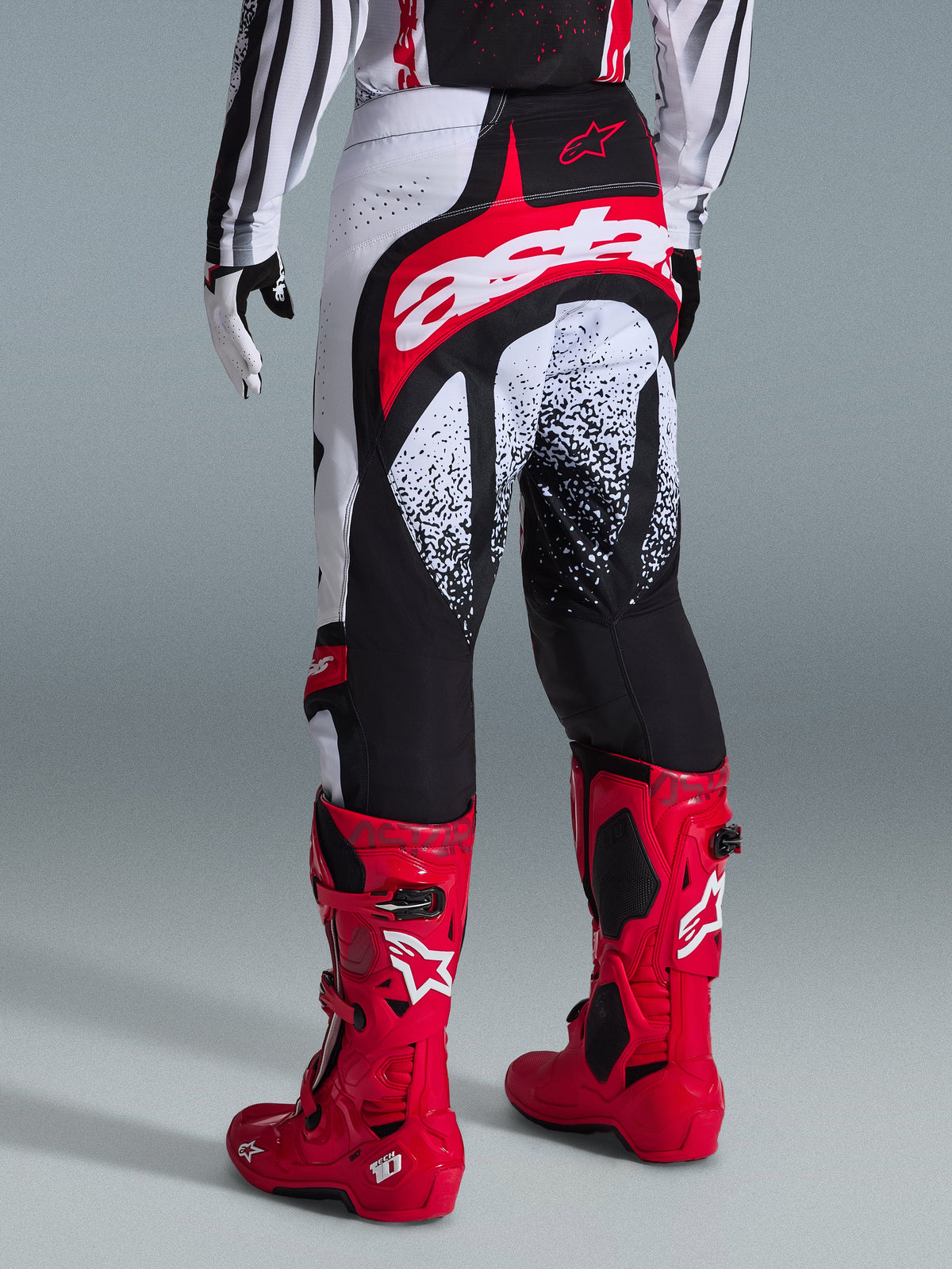 Alpinestars 2026 Techstar Nomur broek, motorcross broek, zwart wit en rood, achteraanzicht getoond op een rijder met bijpassende rode laarzen, voorzien van een opvallend rood en wit 'astars'-logo op het zitvlak en gespikkelde grafische patronen, professionele off-road race-uitrusting