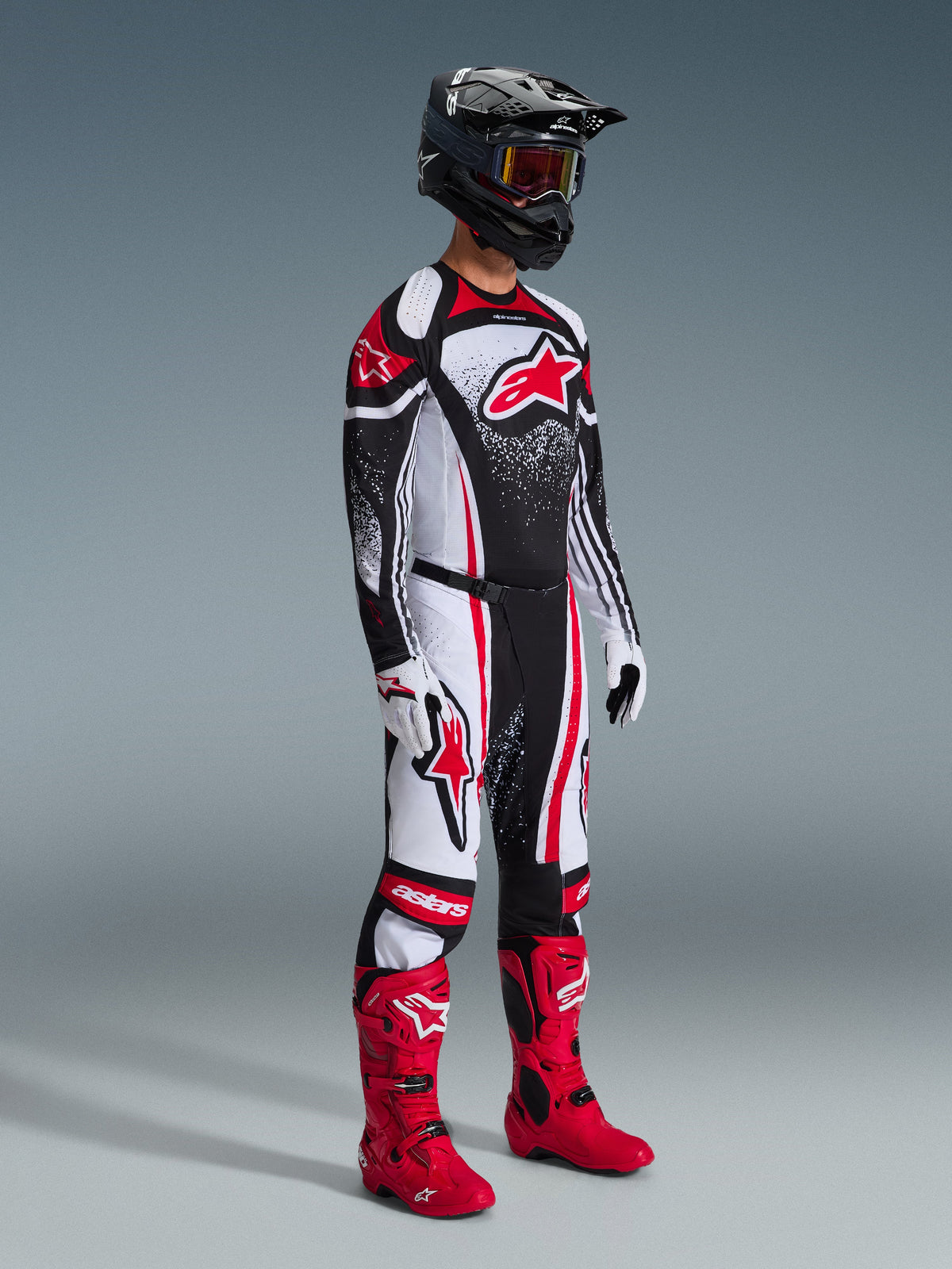 Motocrosser in volledige race-uitrusting, 2026 Techstar Nomur broek in zwart, wit en rood, bijpassende jersey met opvallend Alpinestars-logo en gespikkeld ontwerp, felrode motorcrosslaarzen, witte en zwarte handschoenen en een zwarte helm met crossbril, gecoördineerde professionele off-road outfit