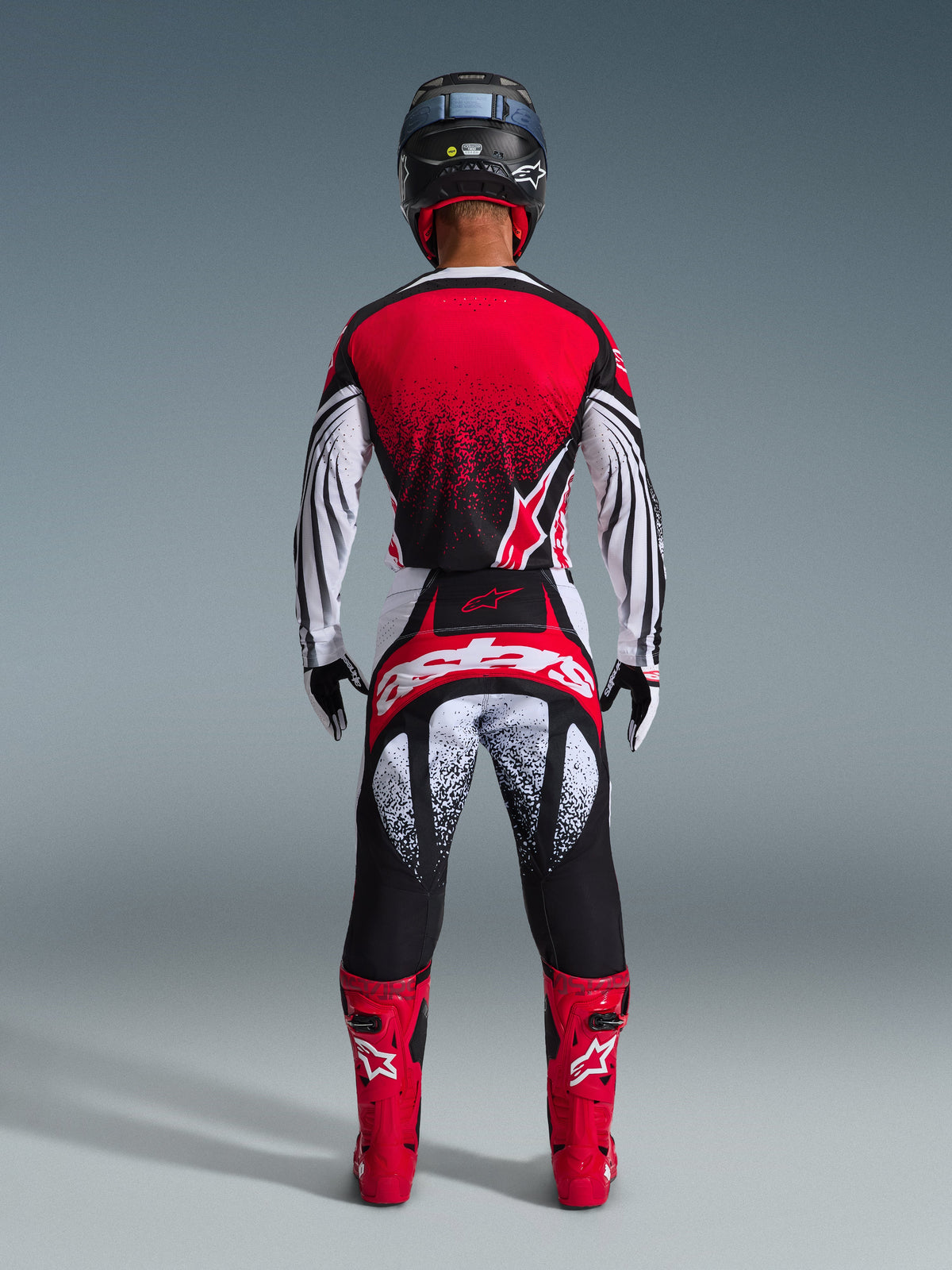 Achteraanzicht van een motorcrosser die de Alpinestars 2026 Techstar Nomur Broek in Black White Red draagt, met opvallende branding op het zitvlak en gespikkelde graphics, gecombineerd met een bijpassende rode jersey en felrode laarzen voor een complete off-road race-uitrusting.