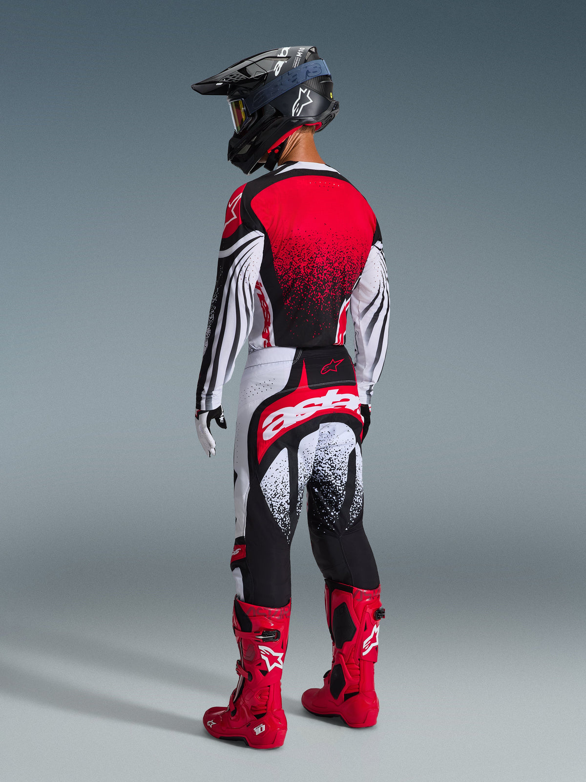 Alpinestars 2026 Techstar Nomur broek, motorcrosser van achteren gezien, zwart wit en rood, met groot wit 'astars' logo op het zitvlak, bijpassende jersey, felrode laarzen en zwarte helm, professionele off-road uitrusting