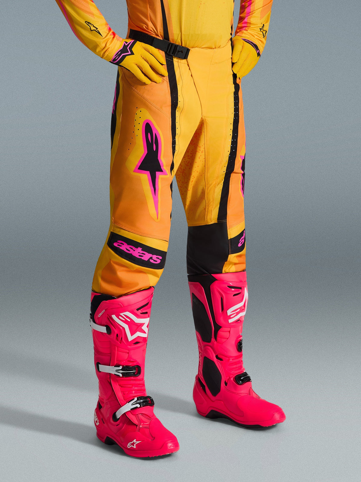 Alpinestars 2026 Techstar Nomur broek, motorcrossbroek, oranje, geel en roze, gedragen met bijpassende gele jersey en handschoenen en felroze laarzen, atletische racepasvorm met roze branding en zwarte magnetische gesp, levendige uitrusting voor off-road competitie