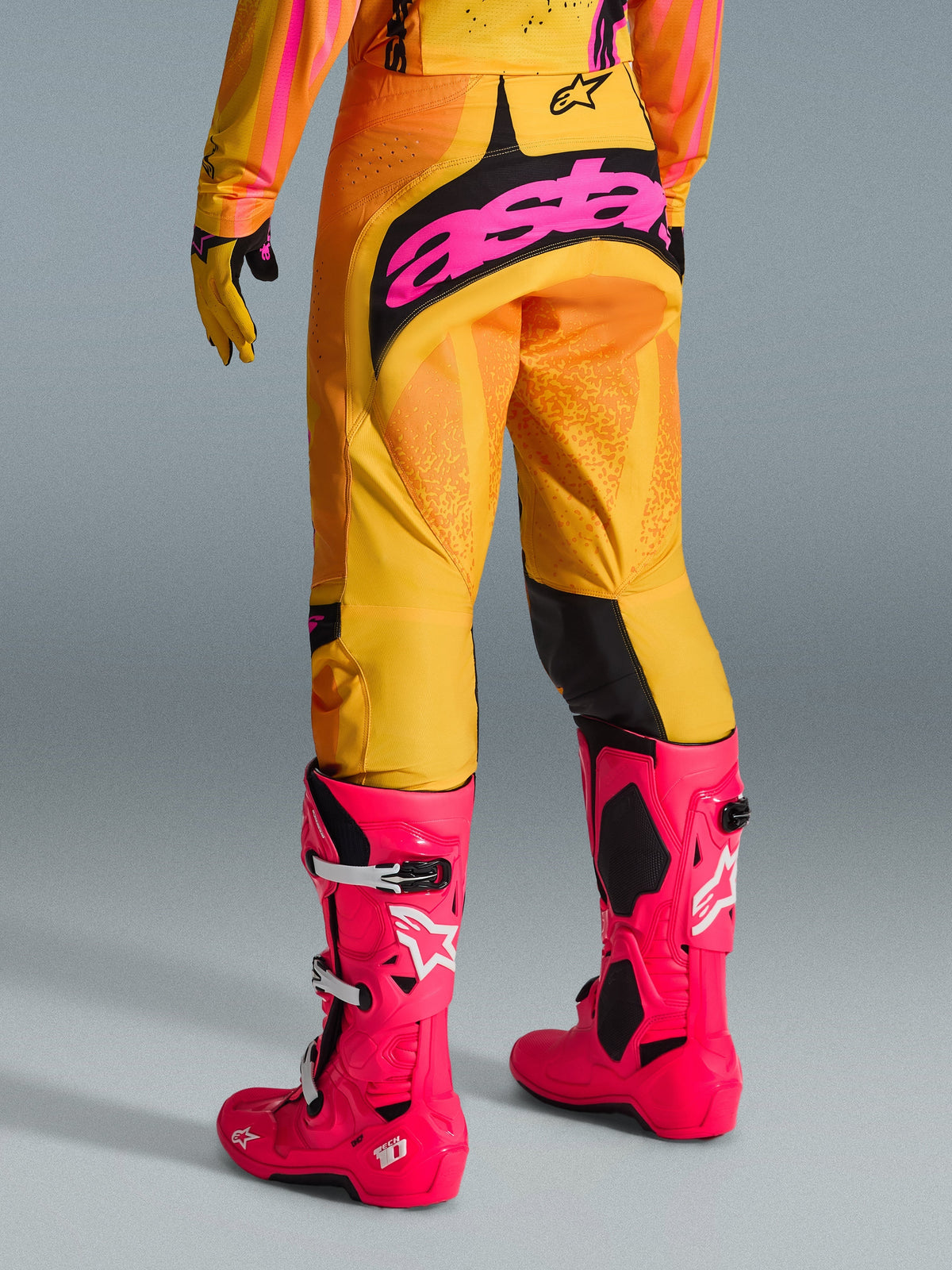 Alpinestars 2026 Techstar Nomur crossbroek, motocrossbroek, oranje, geel en roze, achteraanzicht, met groot roze 'astars' logo op het zitvlak, gecombineerd met felroze Tech 10 laarzen en bijpassende jersey, hoogwaardige race-uitrusting voor motocross