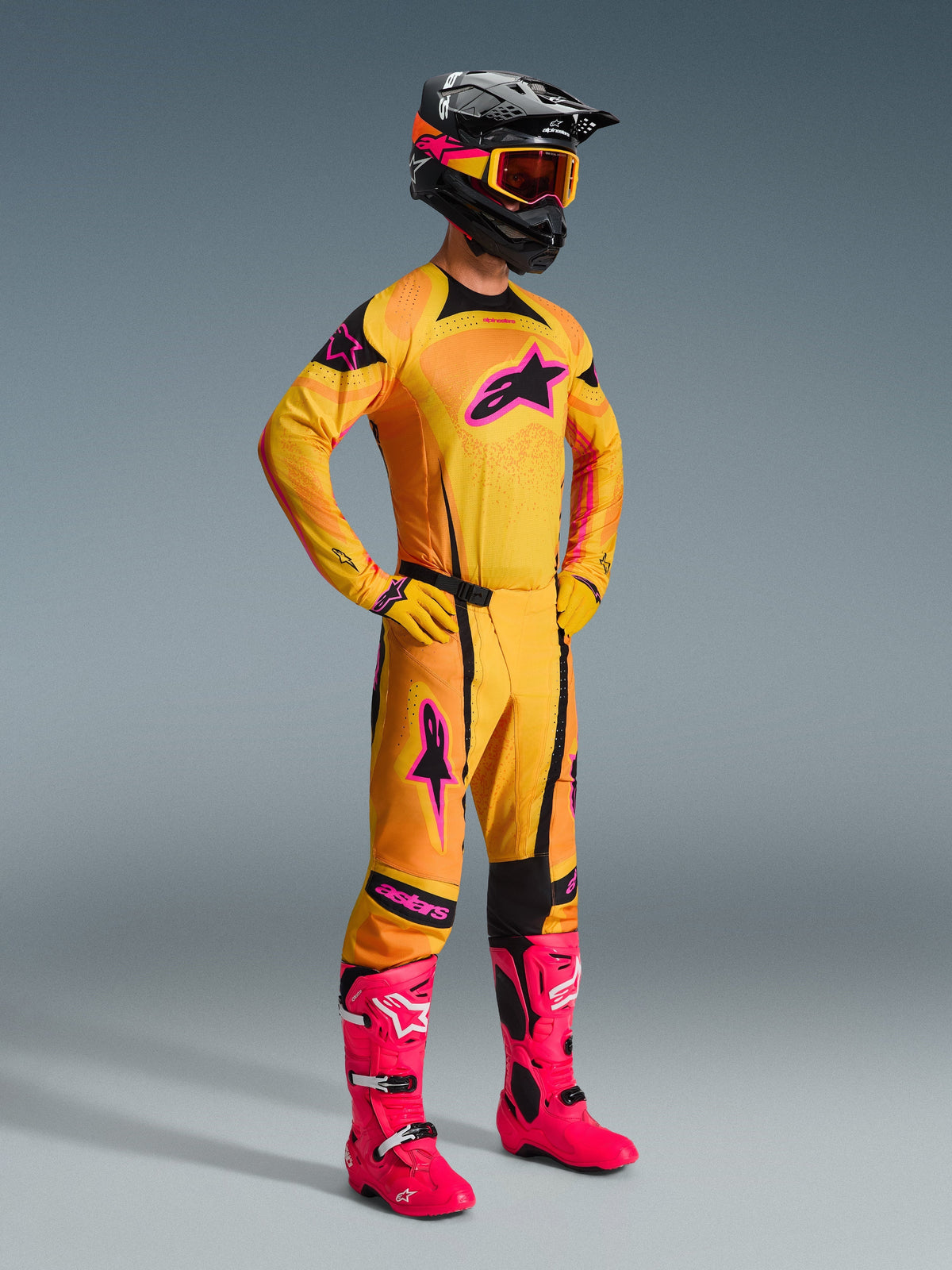 Alpinestars 2026 Techstar Nomur broek in Oranje Geel Roze, complete motorcross-outfit met bijpassend shirt en handschoenen, felroze laarzen, zwarte helm met oranje bril, volledig beeld tegen een grijze achtergrond, professionele off-road race-outfit