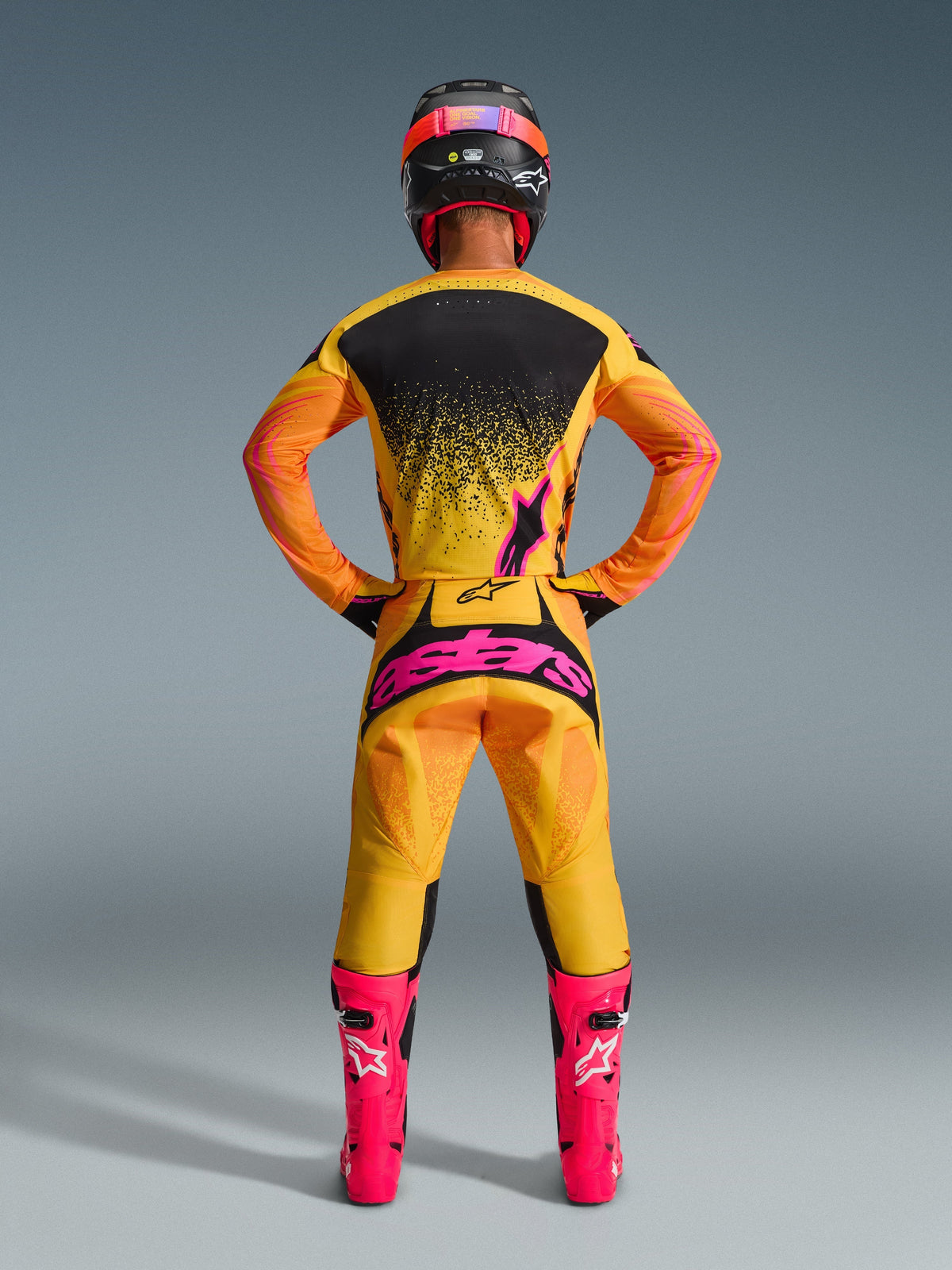 Alpinestars 2026 Techstar Nomur crossbroek, motorcross broek, achteraanzicht van een rijder in volledige uitrusting, kleur Orange Yellow Pink met groot roze logo op het zitvlak, bijpassende geel-zwarte jersey, felroze laarzen en zwart-roze helm