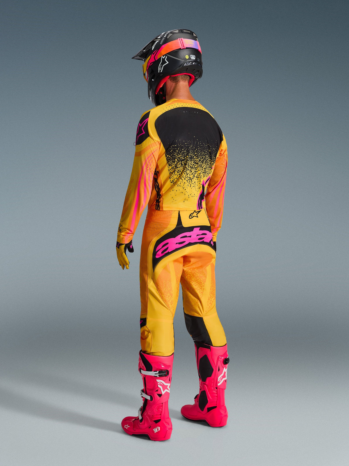 Alpinestars motorcrosser van achteren gezien met complete race-uitrusting, 2026 Techstar Nomur broek in oranje, geel en roze, opvallend roze 'astars' branding op het zitvlak, bijpassende jersey, felroze laarzen en zwarte helm met roze accenten, gecoördineerde off-road wedstrijdoutfit.