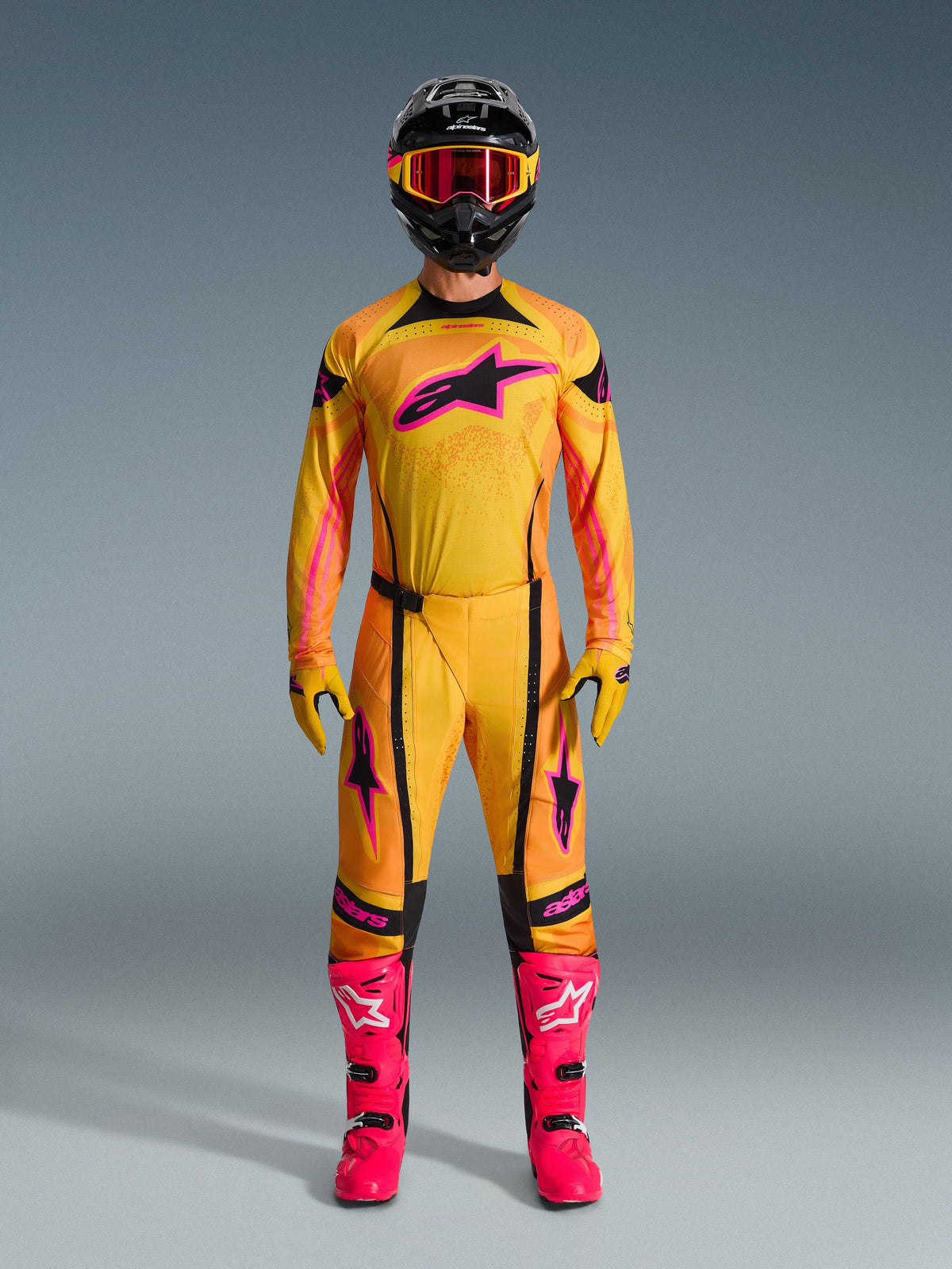 Motorcrossrijder in volledige Alpinestars 2026 Techstar Nomur gearset, oranje, geel en roze jersey en broek, bijpassende gele en roze handschoenen, felle roze racelaarzen en zwarte helm met roze bril, gecoördineerde high-performance motorcross outfit