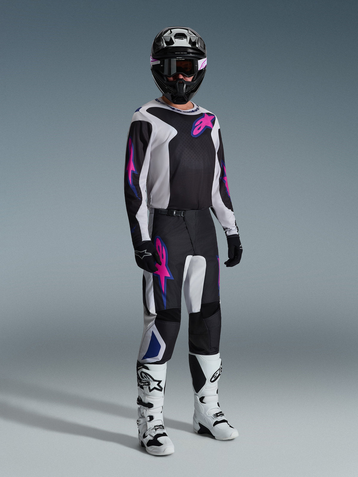 Motocross rider wearing complete racing gear, Alpinestars 2026 Fluid Grid Pants in Black Light Gray and Purple, matching jersey with pink and blue graphics, black helmet and goggles, white and black boots, and black gloves, coordinated outfit for off-road racing