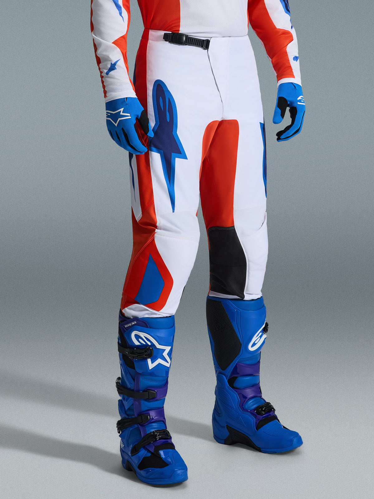 Alpinestars 2026 Fluid Grid Pants, Motocross Pants, Orange and UCLA Blue, worn with matching blue gloves and boots, featuring a white base with bold graphic branding, black inner knee panels, and an ergonomic design for off-road riding.