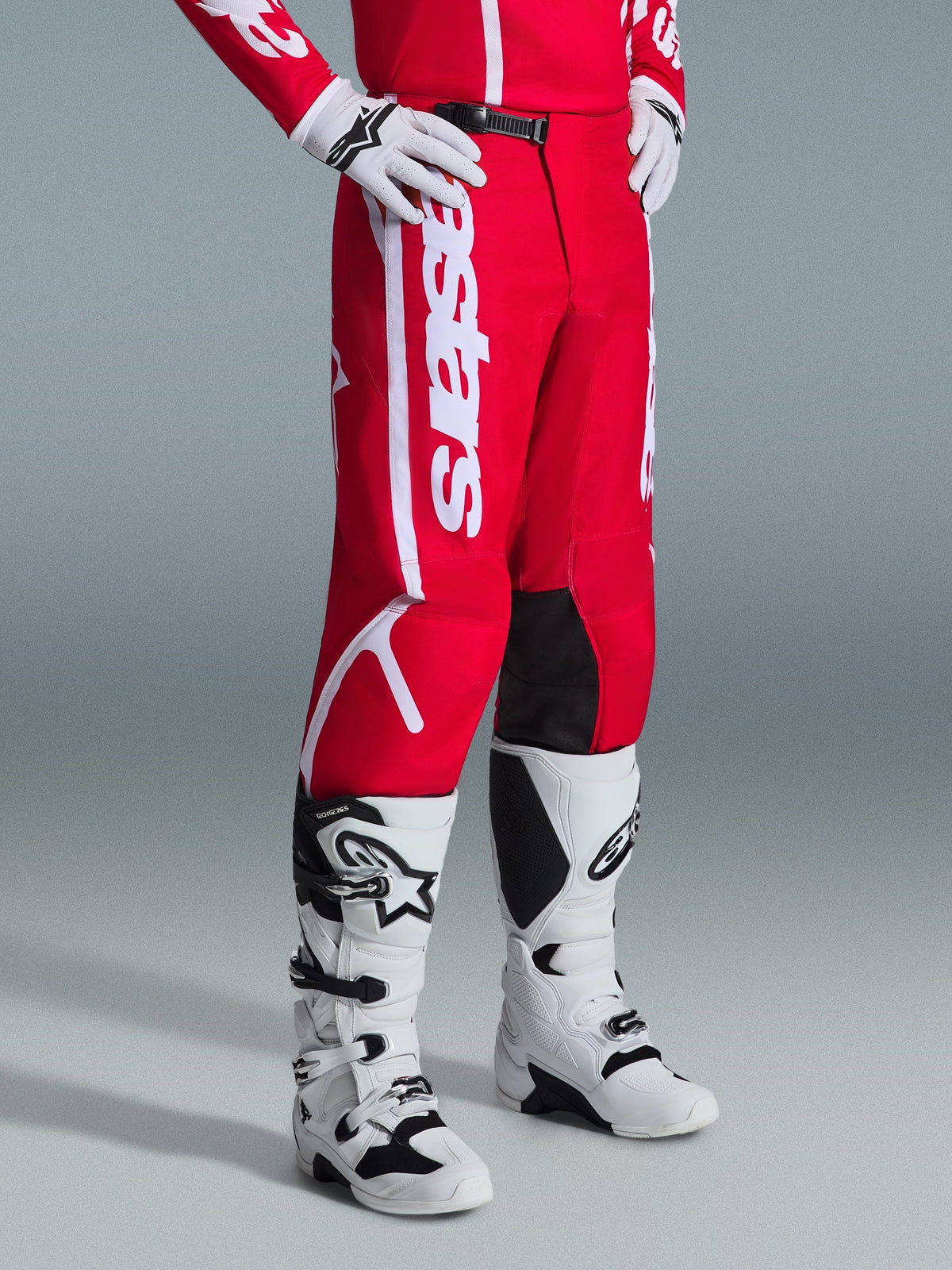 Alpinestars 2026 Fluid Apex Pants, motorcrossbroek, rood en wit, gedragen met witte en zwarte motorcrosslaarzen en handschoenen, voorzien van opvallende witte branding en zwarte panelen aan de binnenkant van de knie, ontworpen voor off-road prestaties