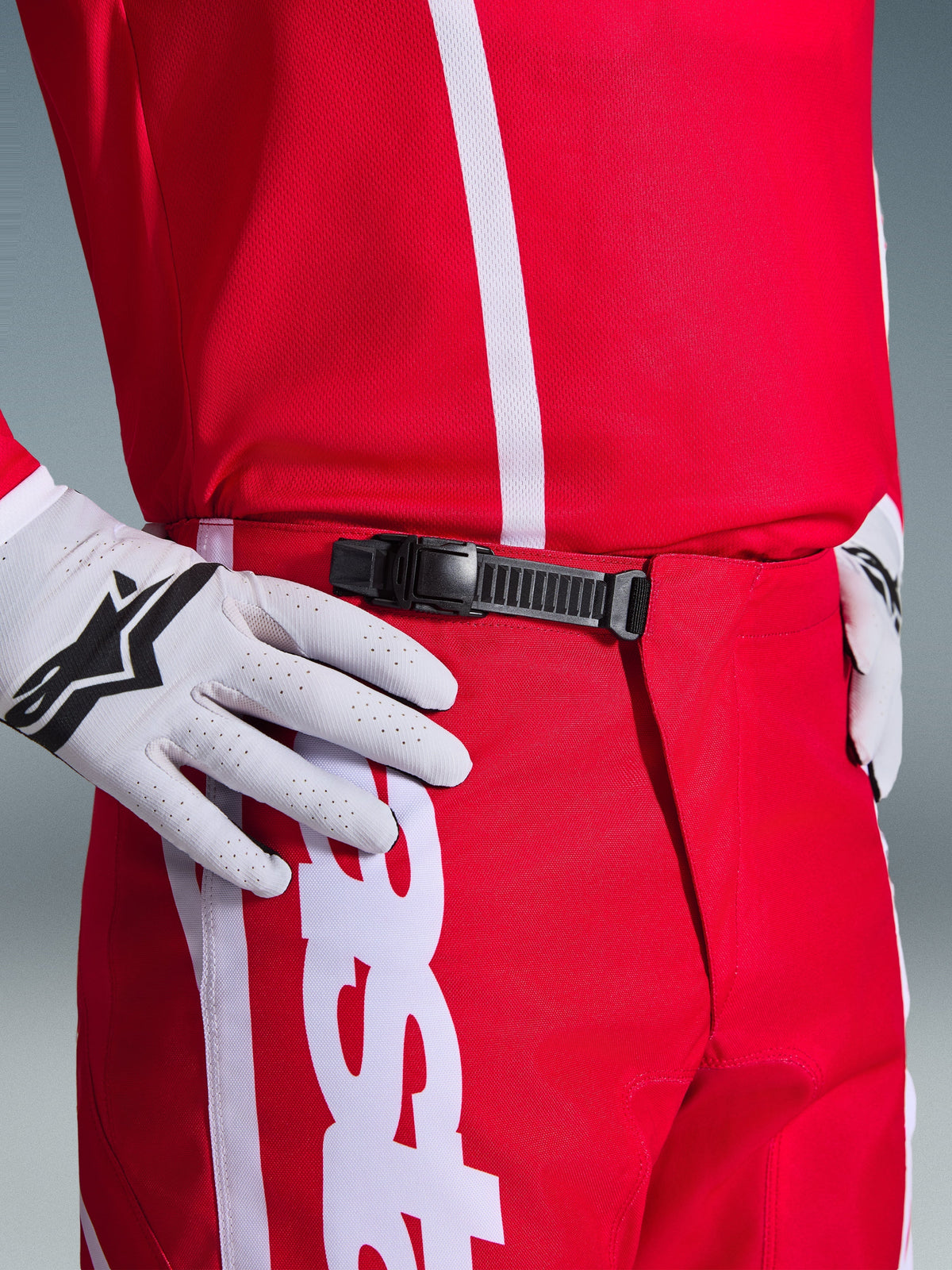 Alpinestars 2026 Fluid Apex broek, motorcrossbroek, rood en wit, close-up van de zwarte ratelsluiting bij de taille, gedragen met witte Alpinestars handschoenen en bijpassend rood shirt, duurzame performance uitrusting voor off-road rijden.