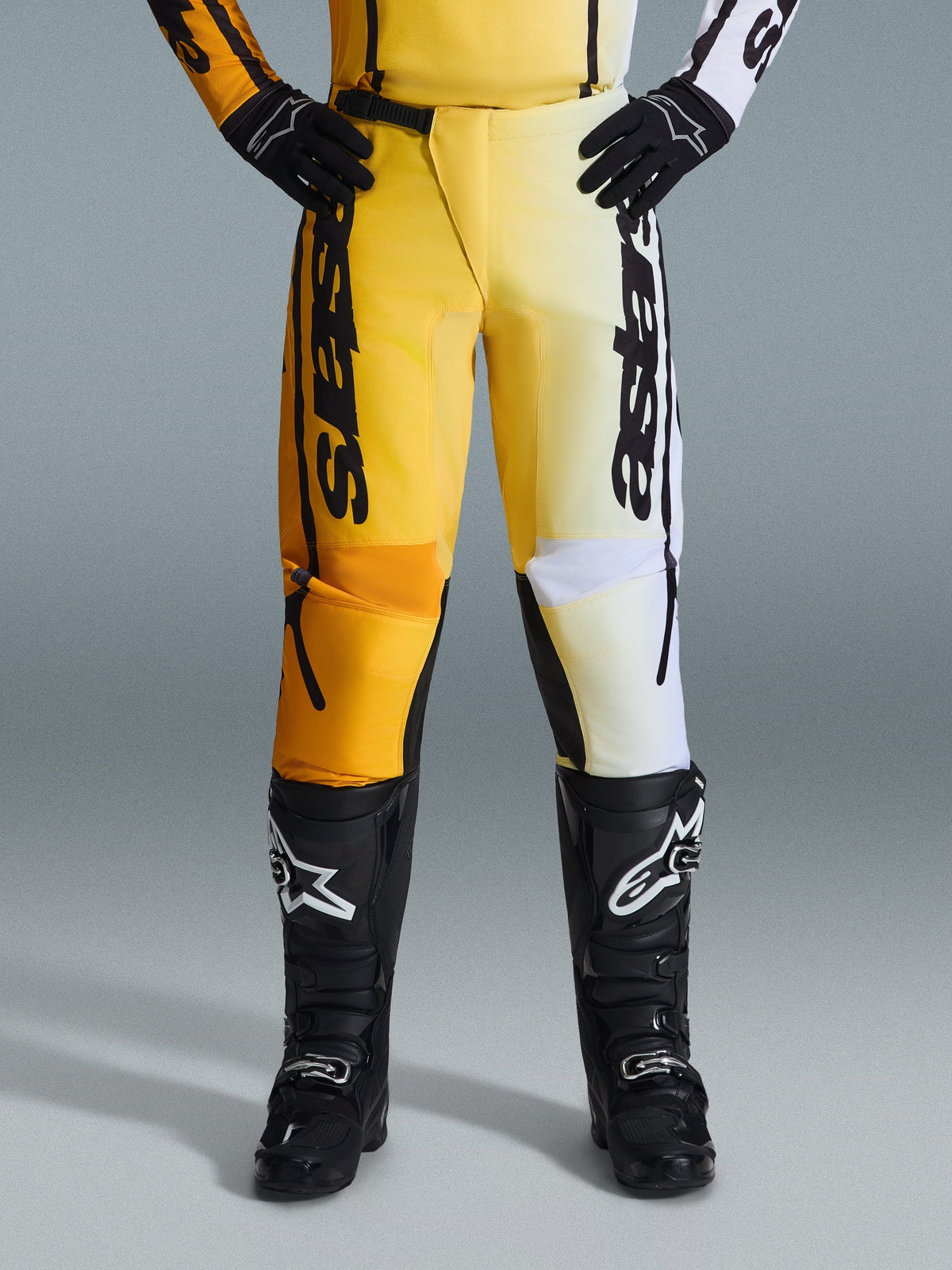 Alpinestars 2026 Fluid Apex broek, motorcrossbroek, geel en zwart, tweekleurig ontwerp met gele en witte panelen, opvallende verticale 'astars' branding, gedragen met zwarte motorcrosslaarzen en handschoenen, performance kleding voor off-road rijden