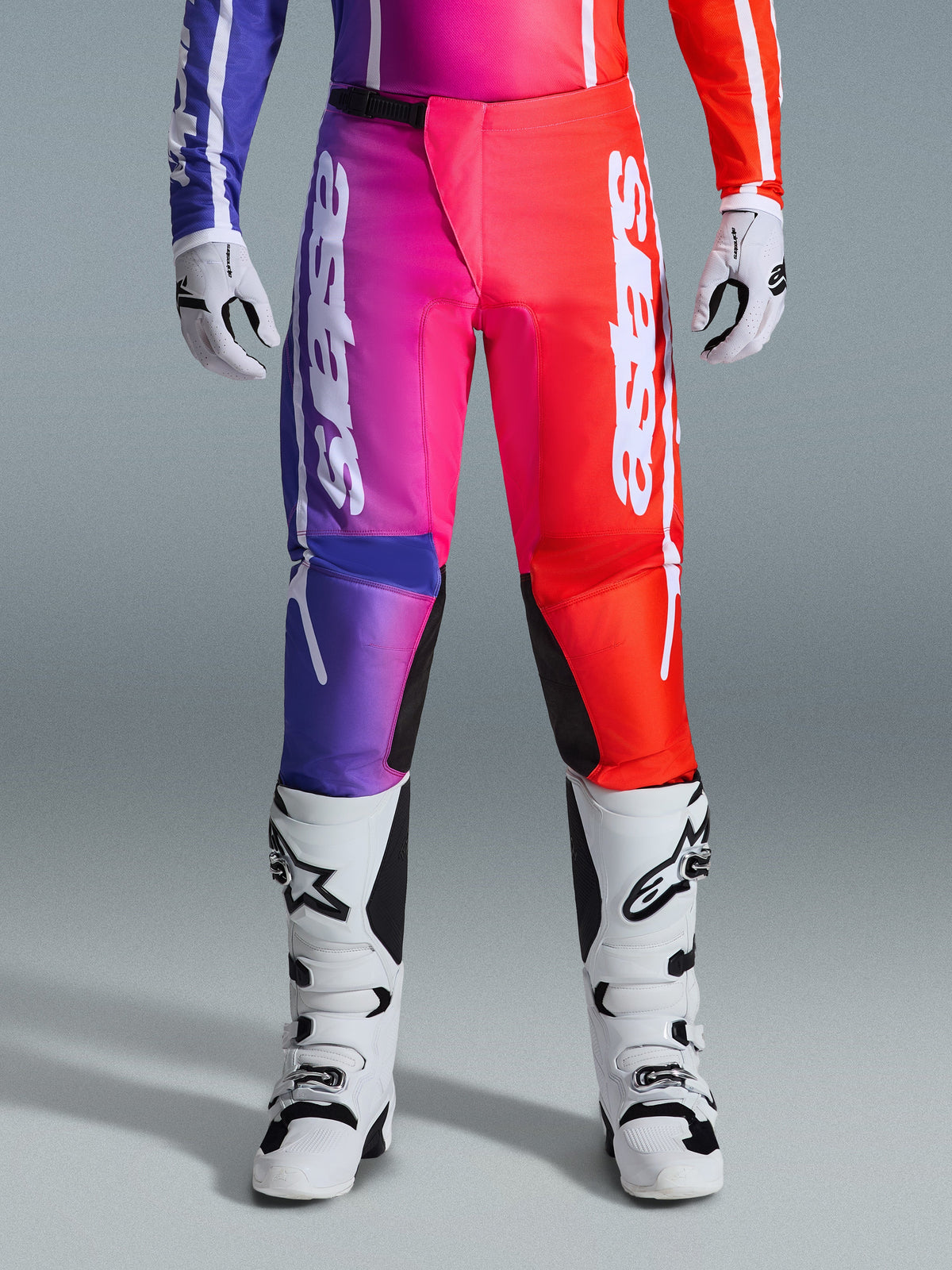 Alpinestars 2026 Fluid Apex Pants, Motocross Pants, Multicolor purple, pink, and red gradient design, large white Alpinestars branding on the legs, paired with white and black motocross boots and gloves, professional off-road racing gear.