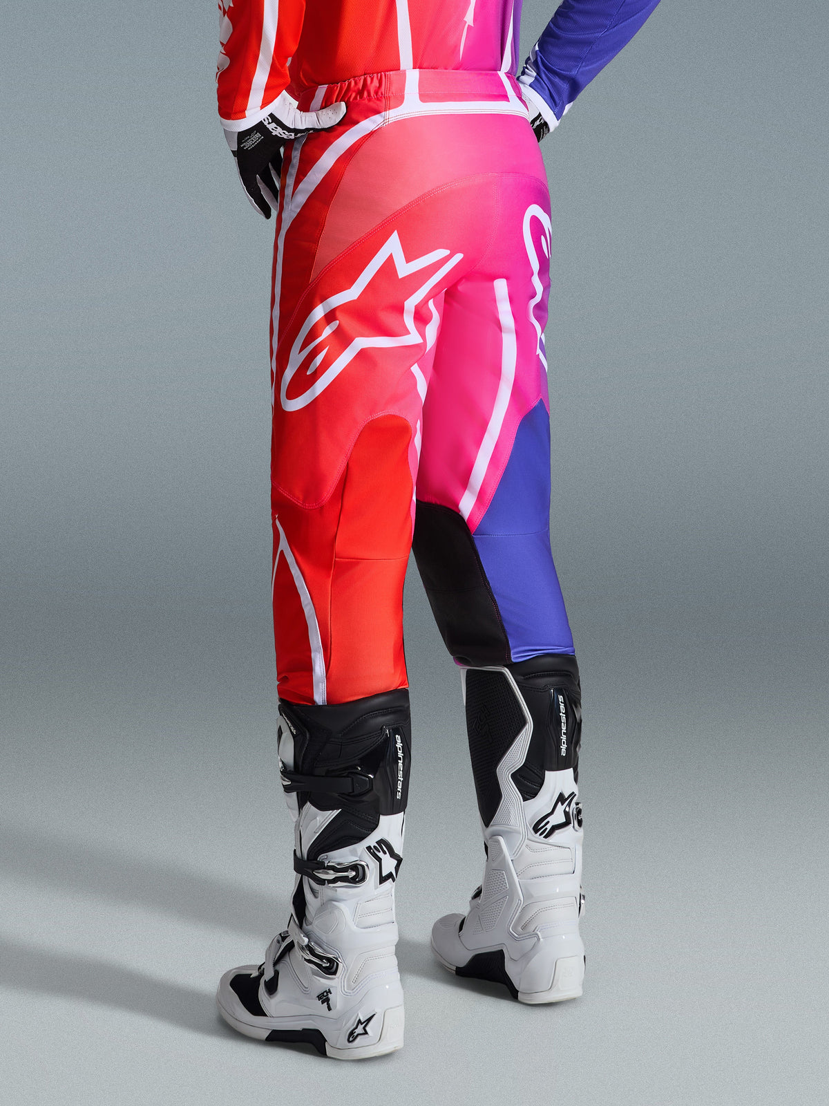 Alpinestars 2026 Fluid Apex Pants, Motocross Pants, Multicolor Red Pink and Purple, Rear view of rider wearing pants with white and black motocross boots, featuring large white Alpinestars logo, vibrant gradient design
