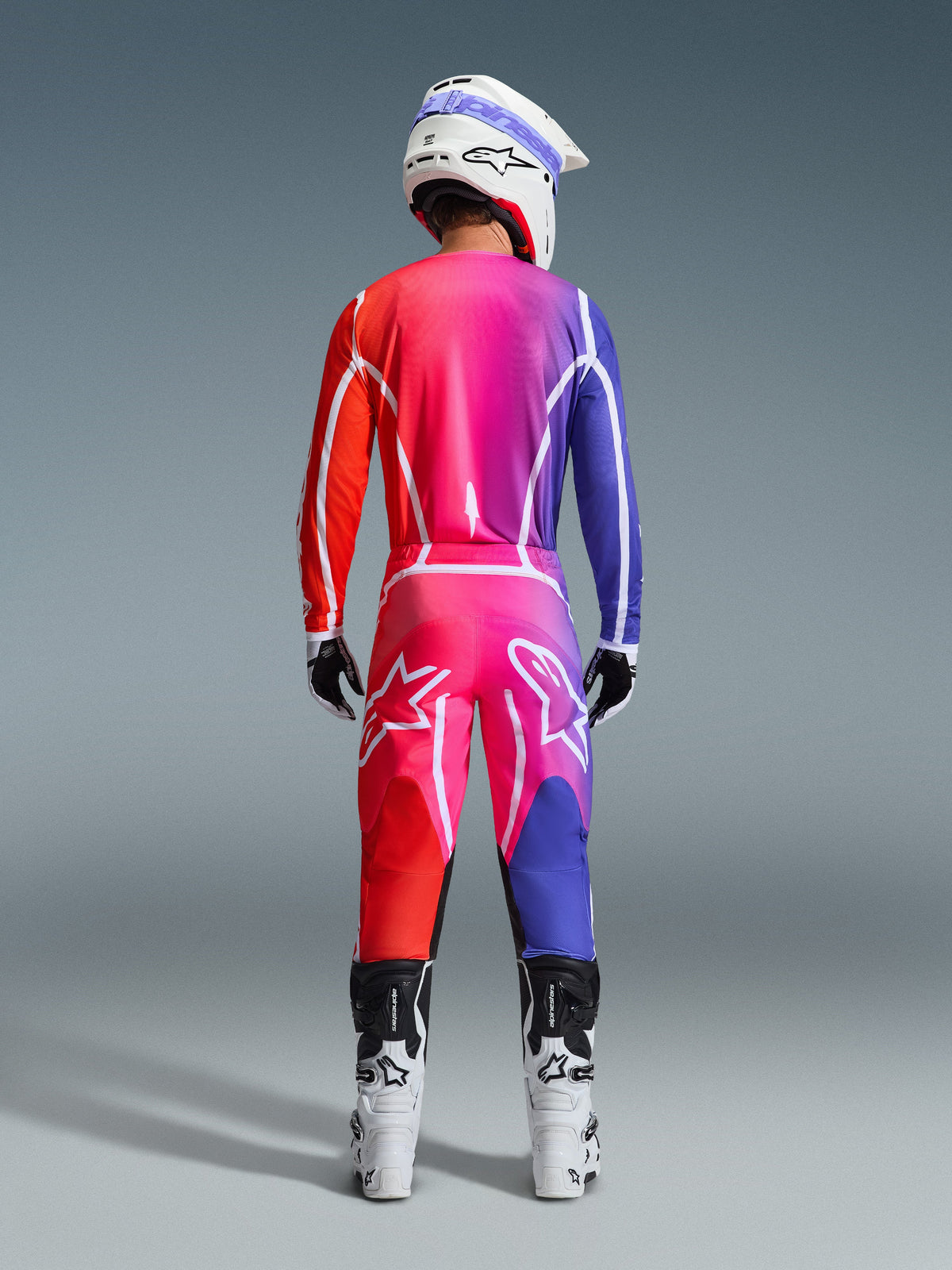 Rear view of a motocross rider wearing Alpinestars 2026 Fluid Apex Pants and matching jersey, featuring a vibrant red, pink, and purple gradient, paired with a white helmet and white boots, complete coordinated gear set for off-road racing
