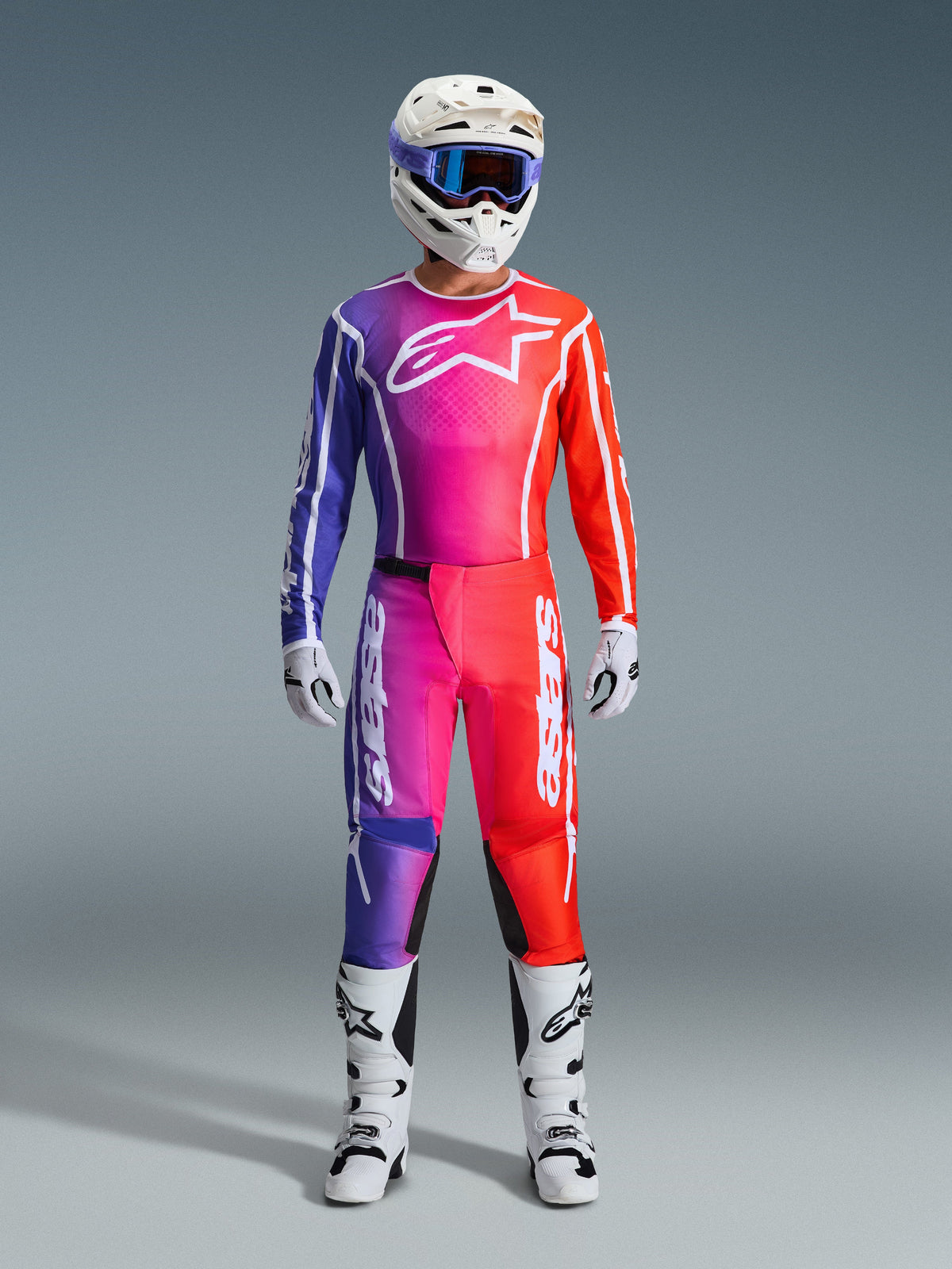 Motocross rider wearing 2026 Fluid Apex Pants and matching jersey, purple and orange gradient, white helmet with goggles, white boots and gloves, coordinated Alpinestars racing gear for off-road performance.