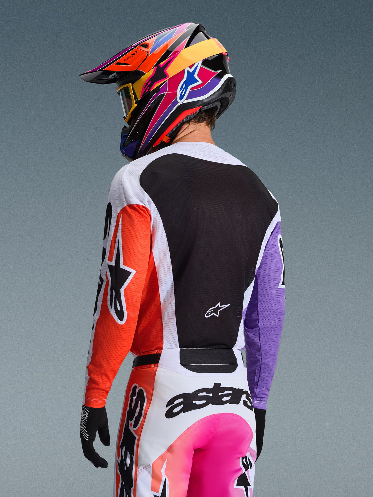 Alpinestars 2026 Racer Air Portl Jersey in Multicolor, motorcrossrijder van achteren gezien, oranje rechtermouw, paarse linkermouw en een groot zwart mesh rugpaneel voor ventilatie, gecombineerd met een bijpassende motorcrossbroek en een kleurrijke helm, professionele off-road race-uitrusting.