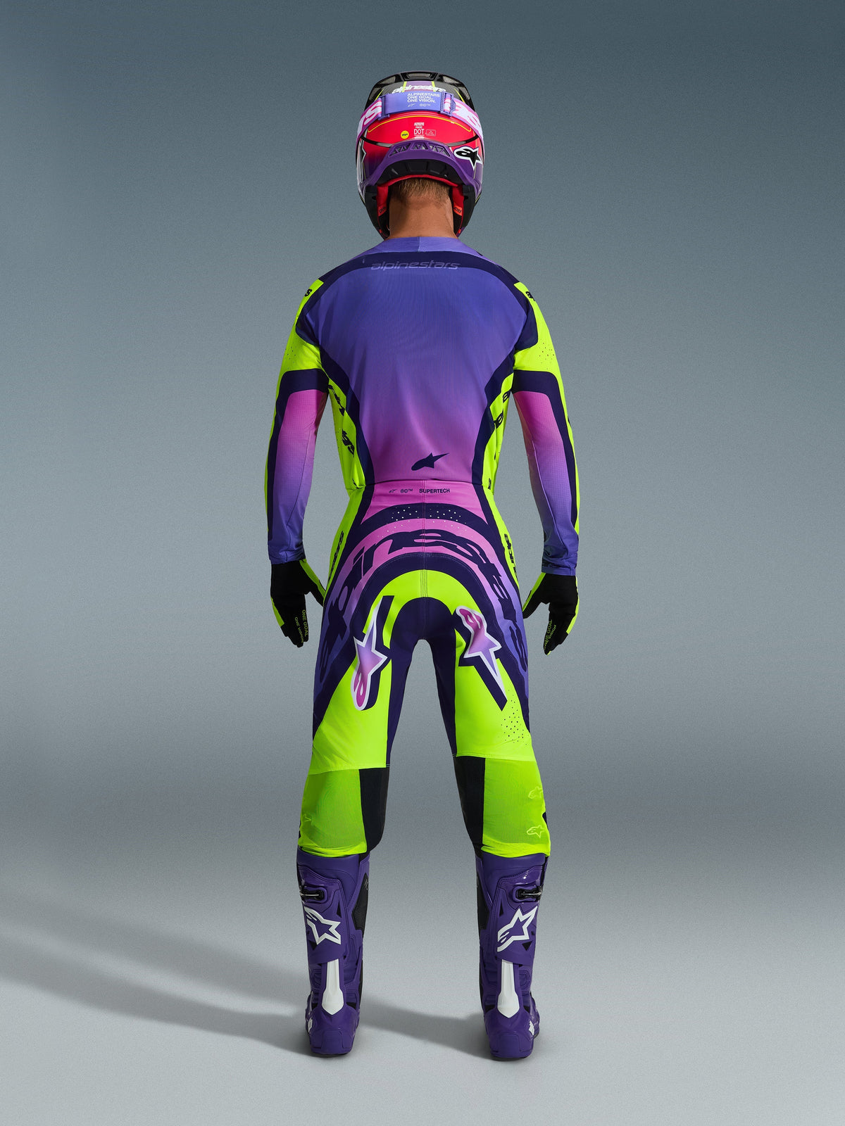 Motocrossrijder van achteren gezien in de 2026 Supertech Vista Jersey en bijpassende broek, opvallend geel en paars ontwerp met roze kleurverlopen, paarse laarzen met witte Alpinestars-logo's, zwarte handschoenen en een rood met paarse racehelm, professionele race-uitrusting set