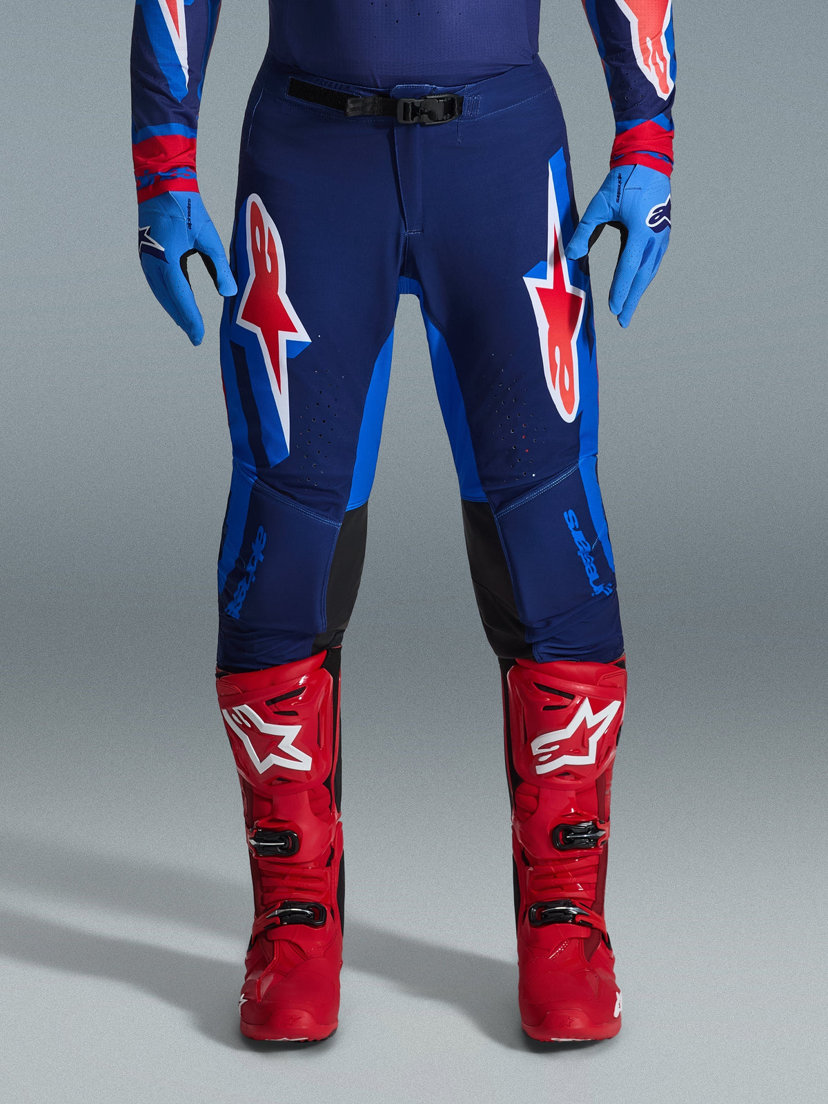 Alpinestars 2026 Supertech Vista broek, motorcrossbroek, donkerblauw, fel koraalrood en kobaltblauw, vooraanzicht van een rijder in racekleding met bijpassende blauwe handschoenen en rode motorcrosslaarzen, met opvallende rode en witte Alpinestars-logo's op de bovenbenen.