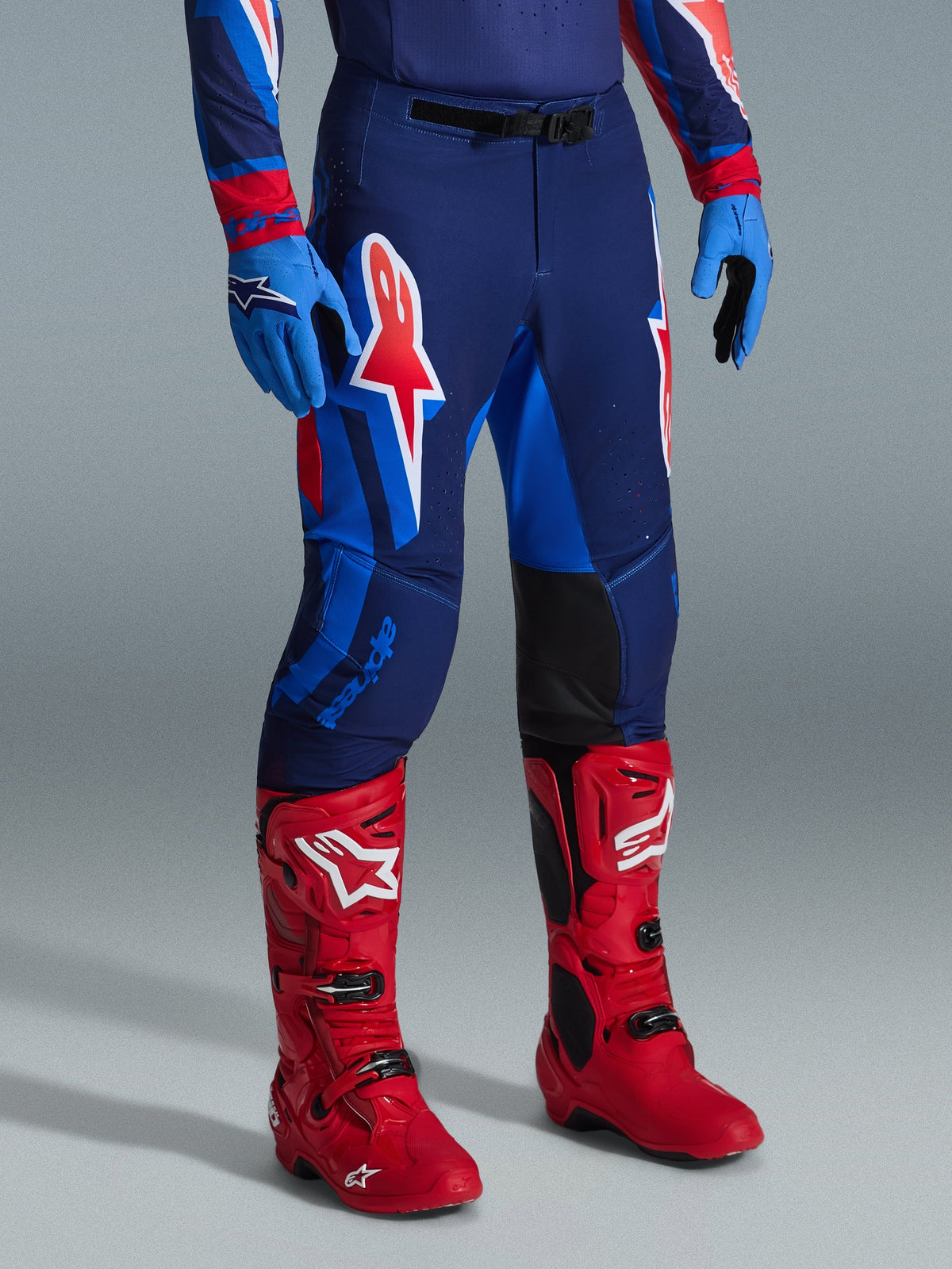 Alpinestars 2026 Supertech Vista broek, motorcross broek, Dark Navy Hot Coral Cobalt, gedragen met felrode racelaarzen en blauwe handschoenen, voorzien van grote grafische logo's op de dijen en zwarte verstevigde binnenkniepanelen voor off-road prestaties.