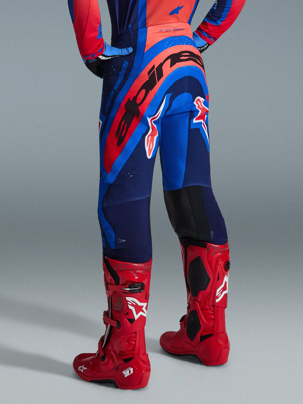 Alpinestars 2026 Supertech Vista broek, motocrossbroek, Dark Navy Hot Coral en Cobalt, achteraanzicht van de rijder, gecombineerd met rode motorcrosslaarzen, opvallende branding op het zitvlak, atletische pasvorm met stretchzones en duurzame rip-stop constructie voor professionele races