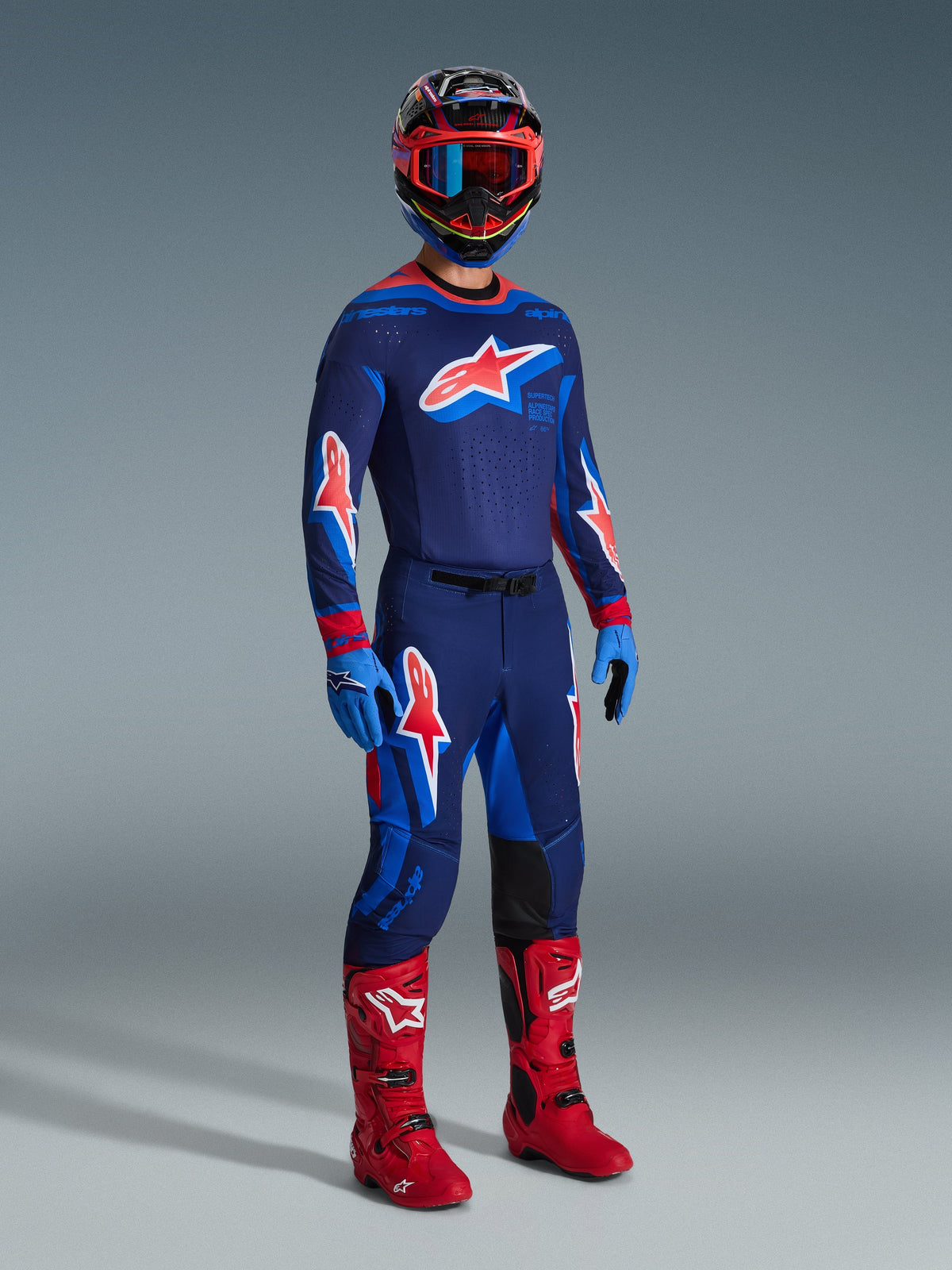 Motorcrossrijder in Alpinestars 2026 Supertech Vista broek in Dark Navy Hot Coral Cobalt, bijpassende racing jersey met lange mouwen, rode motorcrosslaarzen en bijpassende helm met rode bril, volledige studio-opname van hoogwaardige off-road uitrusting