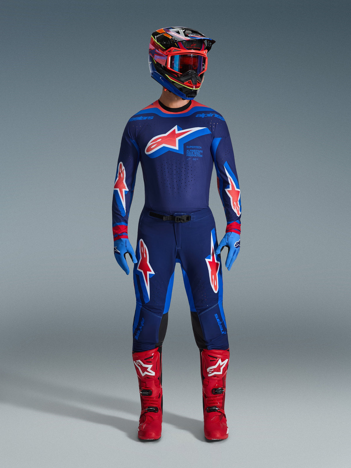 Motorcrossrijder in volledige race-uitrusting, Alpinestars 2026 Supertech Vista broek in Dark Navy Hot Coral Cobalt, bijpassende jersey met opvallend sterlogo, felrode laarzen en helm met rode crossbril, gecoördineerde professionele set voor off-road prestaties