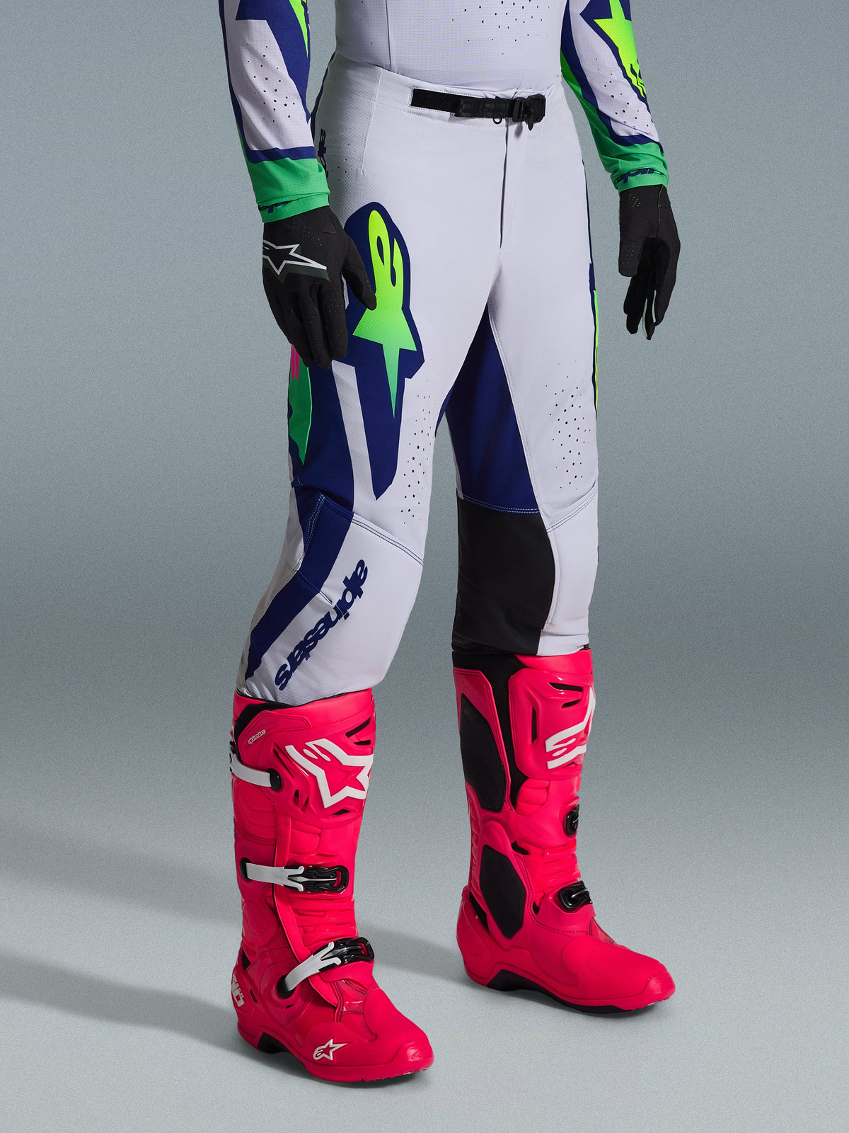 Alpinestars 2026 Supertech Vista Gear Set, motorcrossbroek, lichtgrijs met marineblauwe en limegroene accenten, gedragen met felroze laarzen en zwarte handschoenen, professionele atletische pasvorm en opvallende graphics