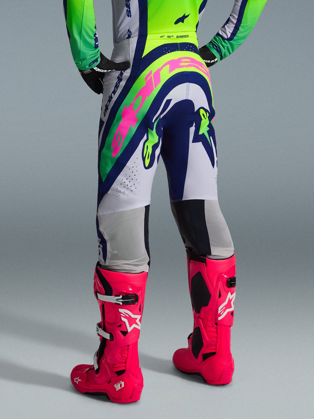 Alpinestars 2026 Supertech Vista broek, motorcrossbroek, lichtgrijs, geel fluo en blauw, achteraanzicht met groot roze Alpinestars-logo, gedragen met bijpassende jersey en felroze Tech 10-laarzen, professionele off-road uitrusting