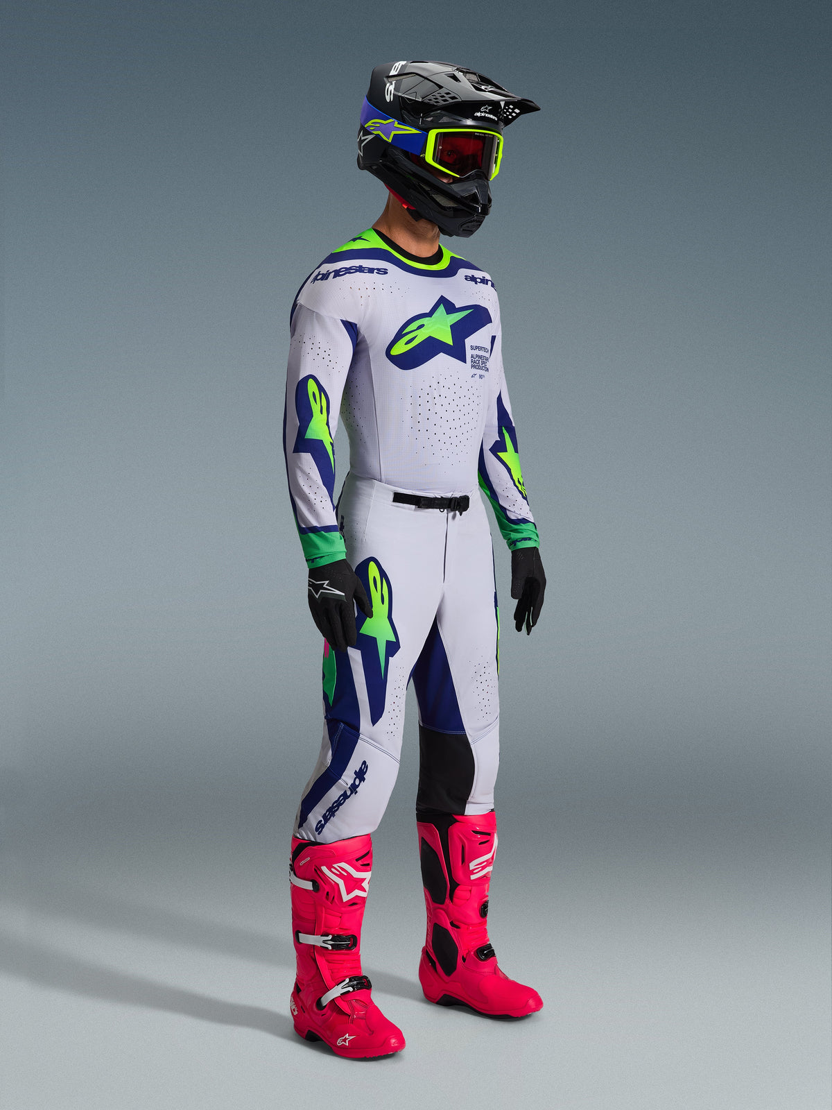 Alpinestars 2026 Supertech Vista Gear Set, motorcrossrijder in volledig racetenue, witte jersey en broek met limoengroene en marineblauwe accenten, felroze laarzen, zwarte helm met blauwe en limoengroene details, gecoördineerde professionele off-road kleding