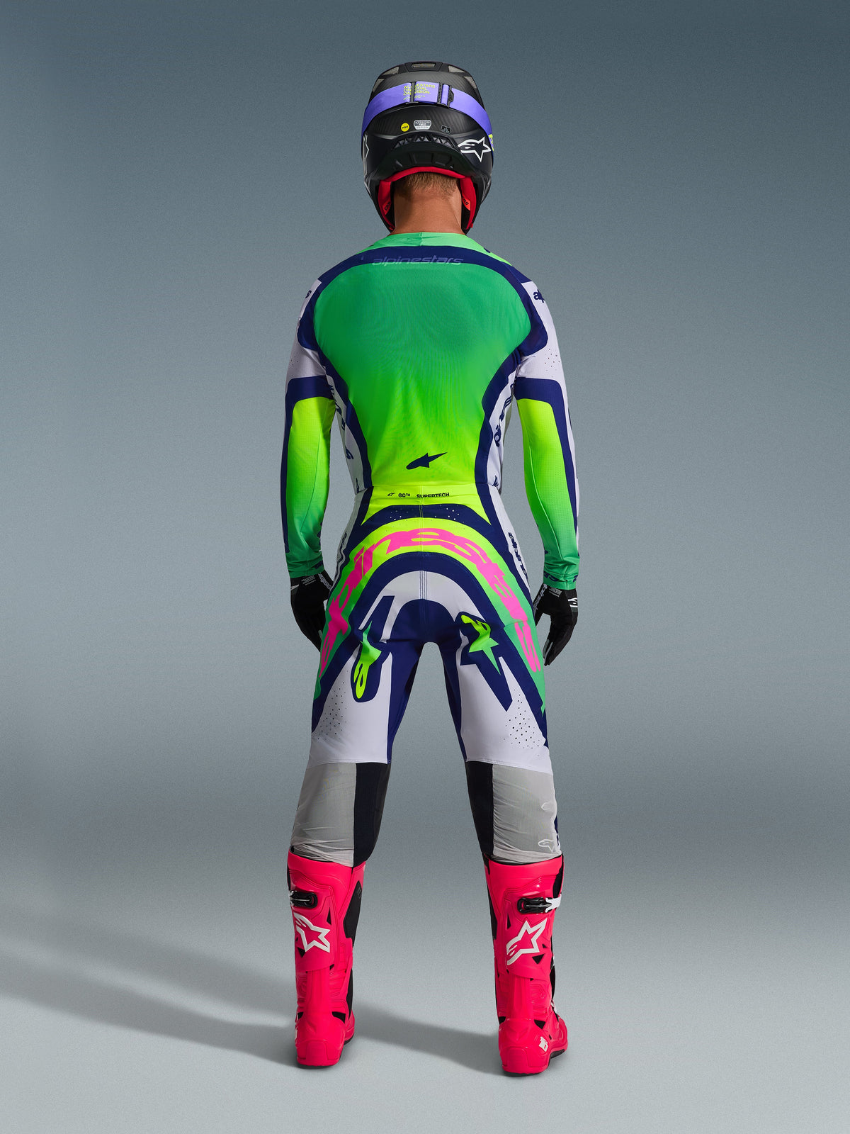 Alpinestars 2026 Supertech Vista Gear Set, motorcross kledingset achteraanzicht, jersey met groen naar geel kleurverloop, witte en blauwe broek met een groot roze logo op de achterzijde, felroze laarzen en een paarse helm, professionele off-road racekleding