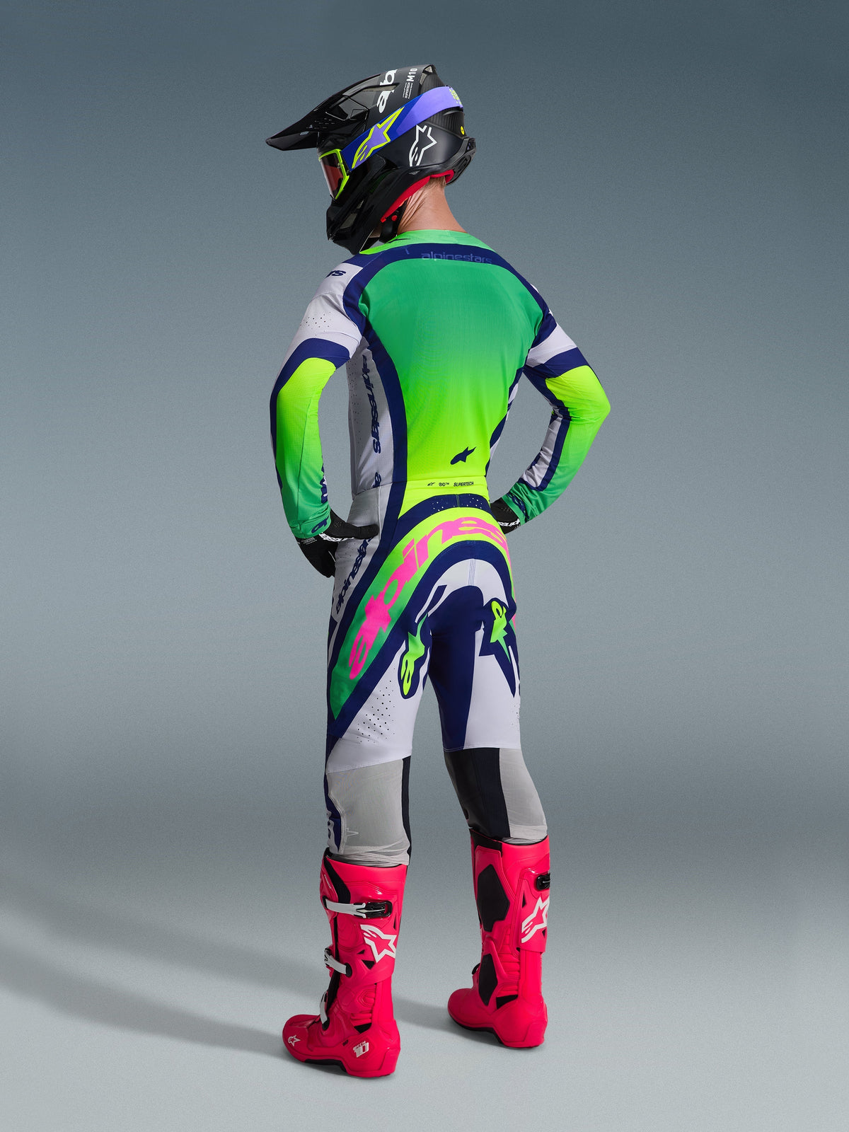 Motorcrosser van achteren gezien in de Alpinestars 2026 Supertech Vista broek, lichtgrijs, blauw en neongeel ontwerp met een opvallend roze logo op het zitvlak, gecombineerd met een bijpassende jersey, zwarte helm en felroze laarzen voor een complete off-road race-uitrusting.