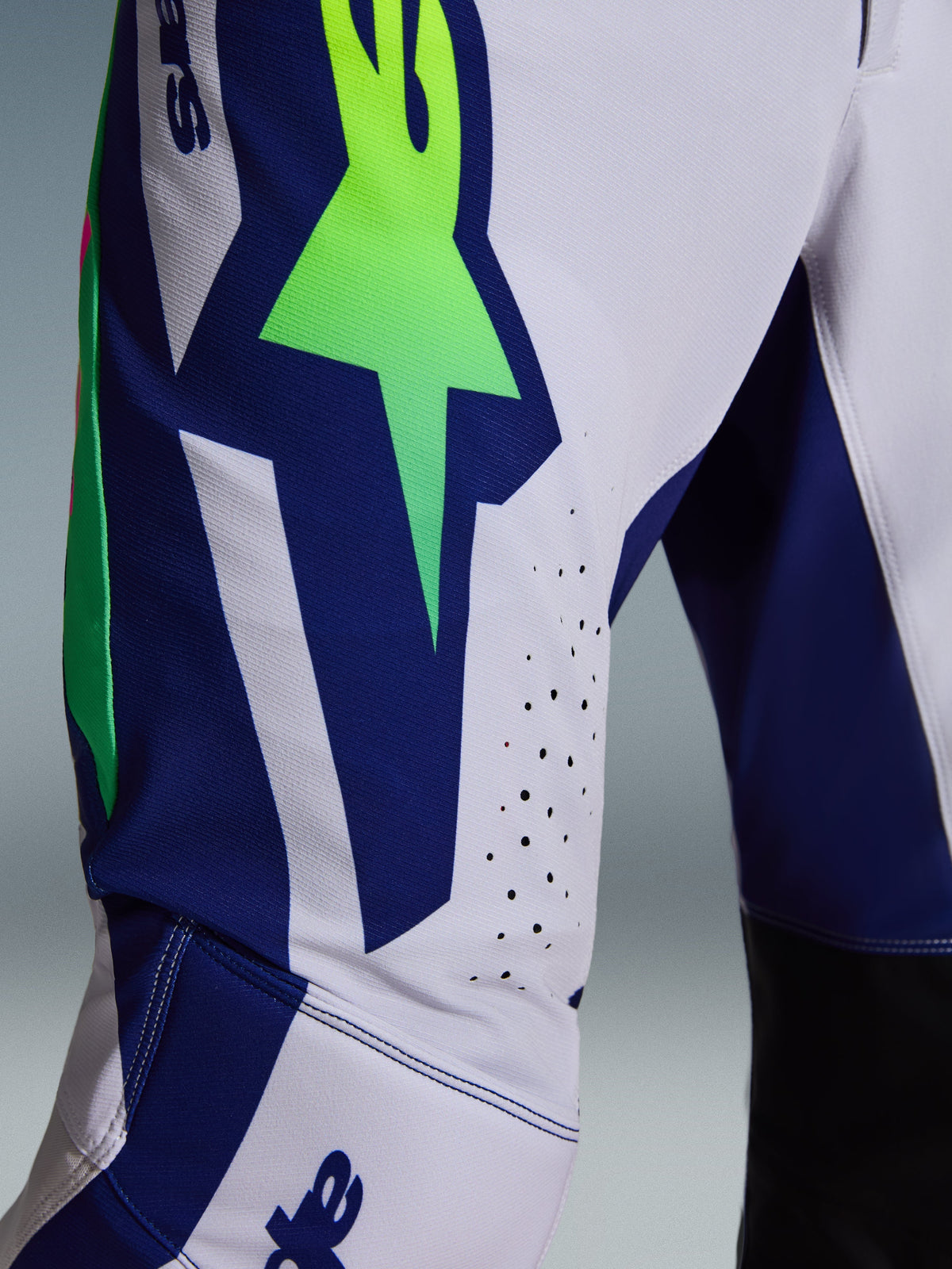 Alpinestars 2026 Supertech Vista Pants, motorcross broek, lichtgrijs, fluorgeel en blauw, detailopname van het dij- en kniegebied met een opvallend neongroen sterlogo op een blauw paneel, lasergesneden perforaties voor ventilatie en zwarte binnenpanelen bij de knie.