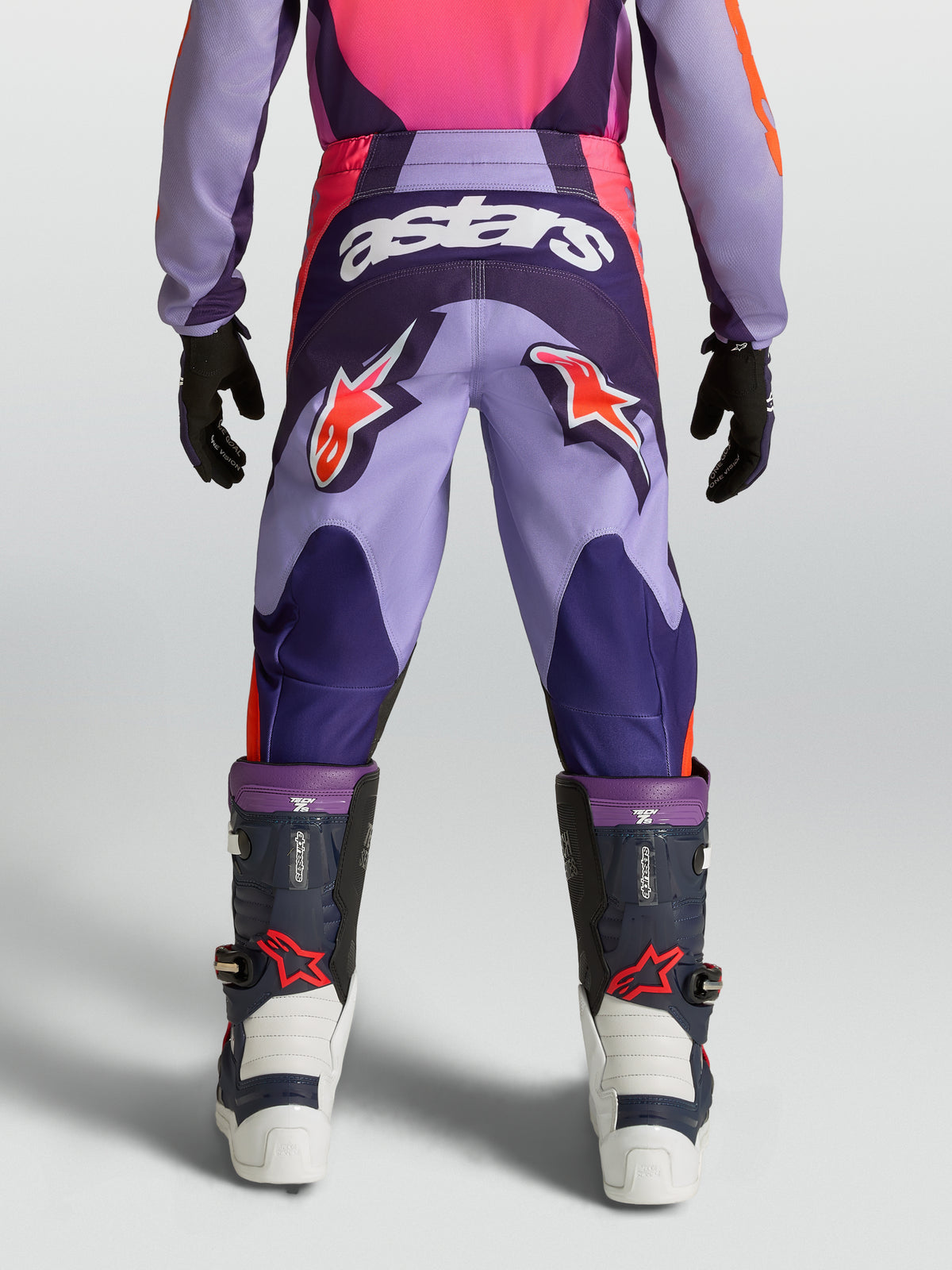 Alpinestars Jeugd Fluid Hunt broek, motocrossbroek voor off-road rijden, paars en roze, duurzame polystof constructie met stretchpanelen, model van achteren gezien met motorlaarzen.