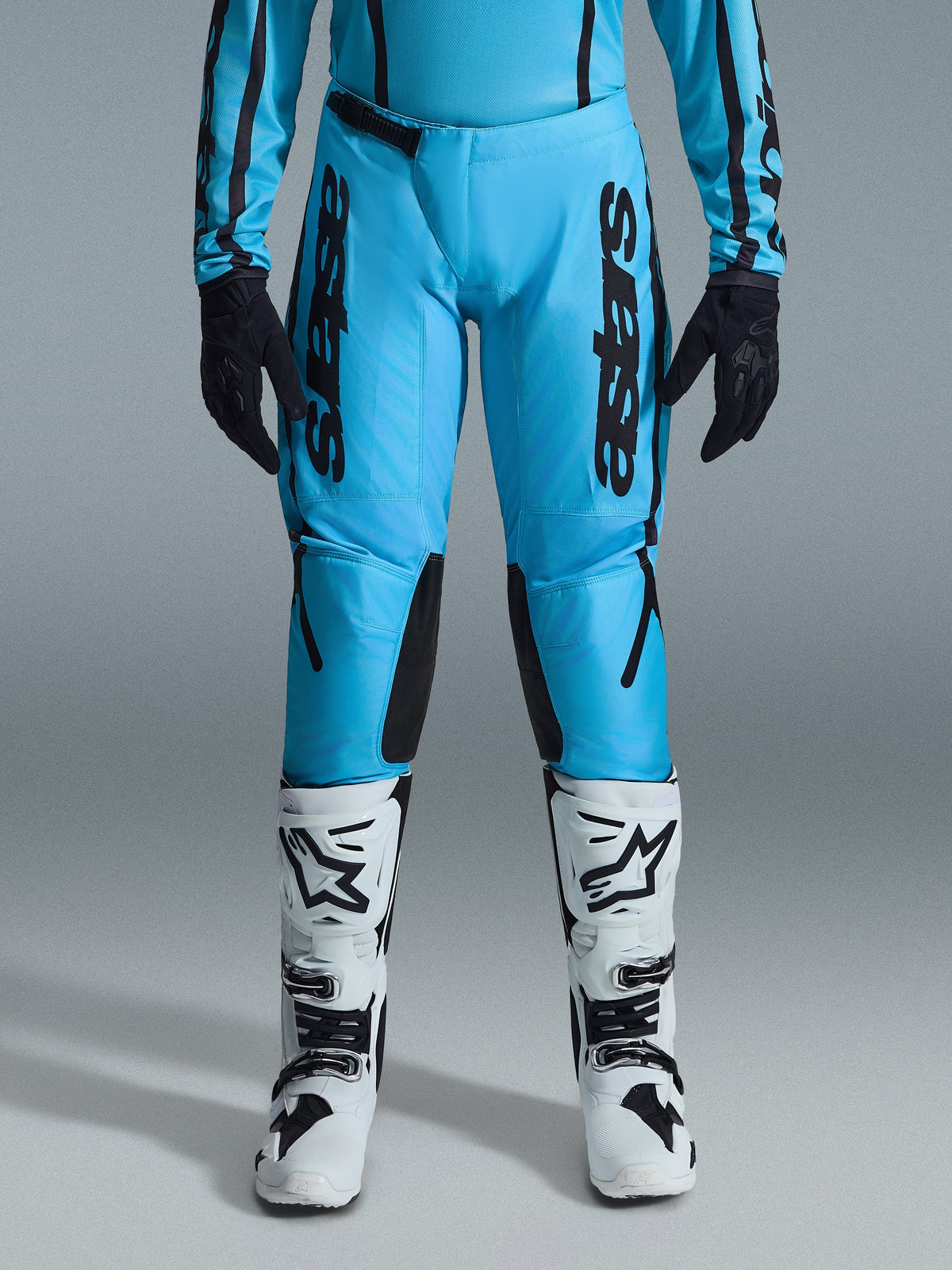 Alpinestars 2026 Stella Fluid Apex damesbroek, motorcrossbroek, fel cyaanblauw en zwart, verticale zwarte branding op de dijen, zwarte verstevigde kniepanelen aan de binnenzijde, getoond met witte en zwarte motorcrosslaarzen en zwarte handschoenen, professionele off-road uitrusting voor dames