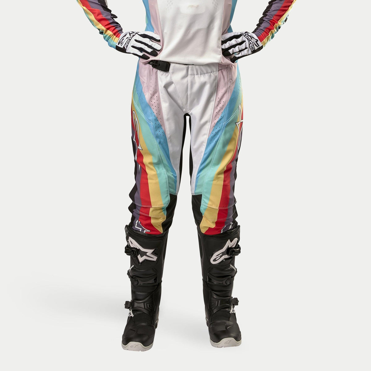 Alpinestars Stella 2024 Techstar dames crossbroek, motorcross broek, zwart multicolor met levendige diagonale strepen in wit, blauw, rood en geel, gedragen met zwarte motorcrosslaarzen en handschoenen, race-geïnspireerd ontwerp voor off-road prestaties.