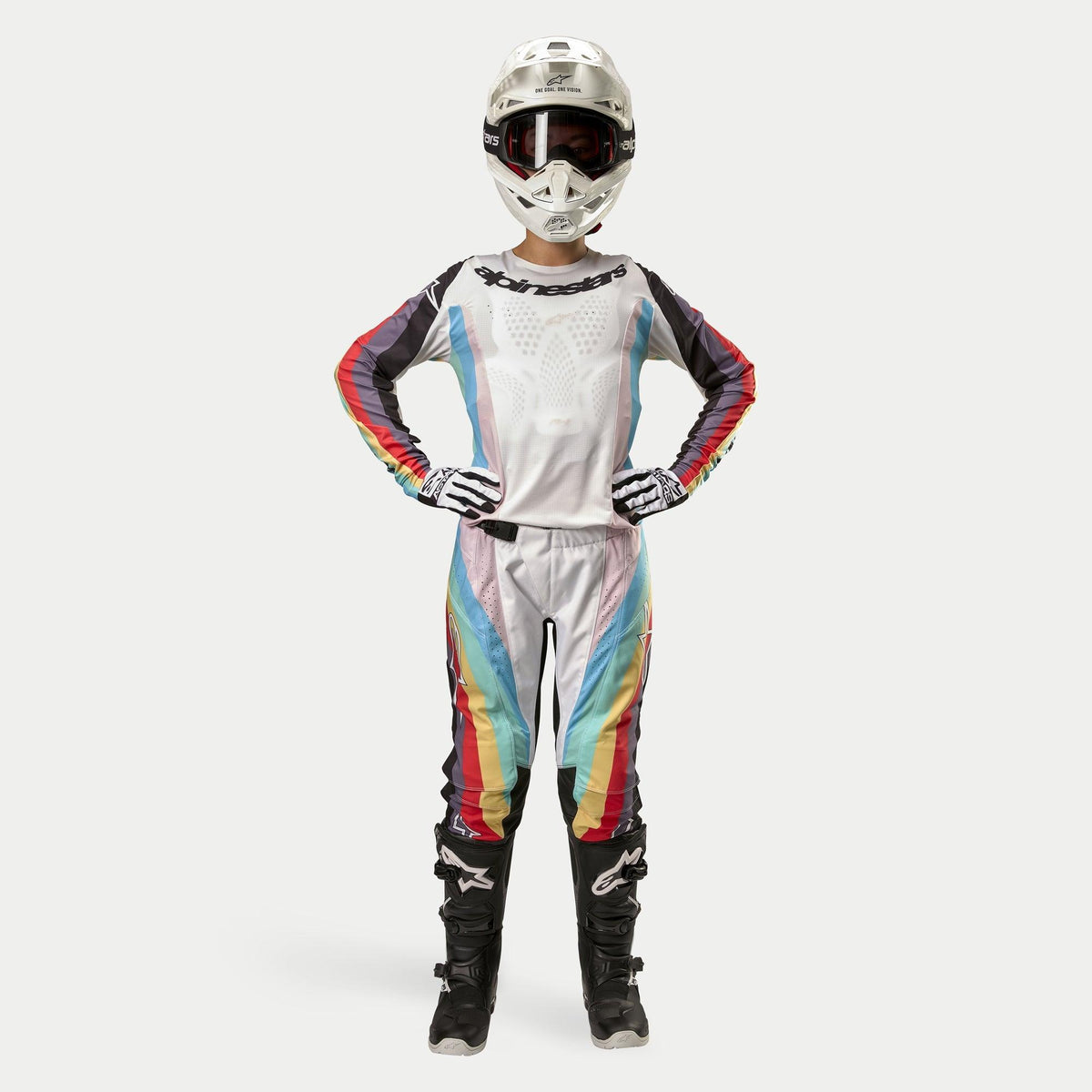Vrouwelijke motorcrosser in Alpinestars Stella 2024 Techstar broek, Black Multicolor, witte basis met rode, gele en blauwe strepen, bijpassende jersey met lange mouwen, witte helm en zwarte laarzen, volledige race-uitrusting voor off-road competitie