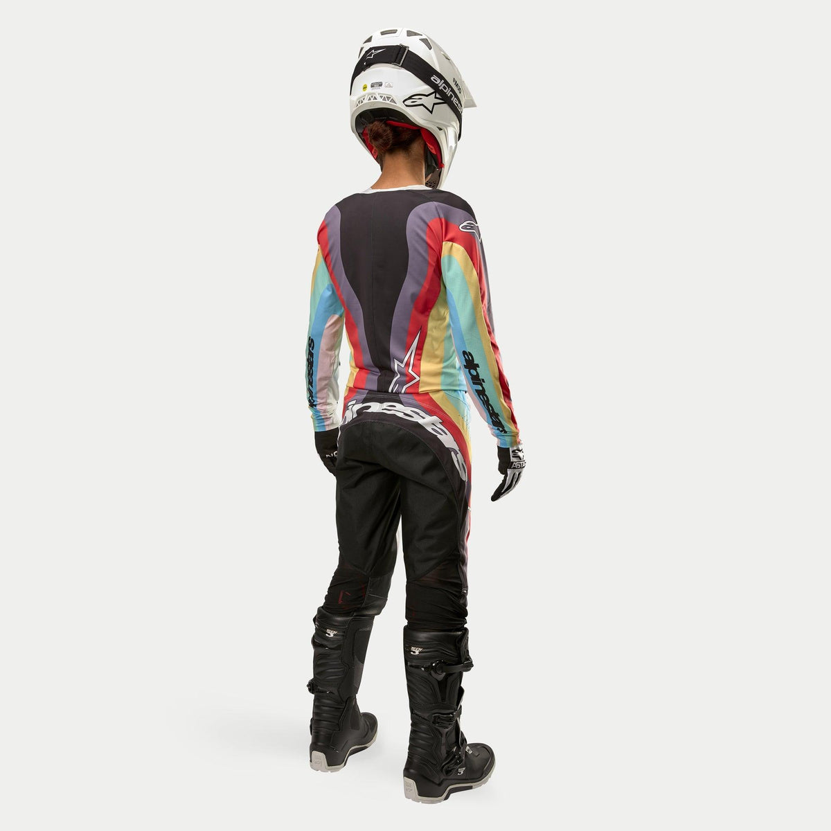 Motorcrosser van achteren gezien in Alpinestars Stella 2024 Techstar damesbroek in Black Multicolor, voorzien van een groot wit logo op het zitvlak, gecombineerd met een kleurrijke gestreepte jersey, witte helm en zwarte Tech 3-laarzen voor off-road racing.