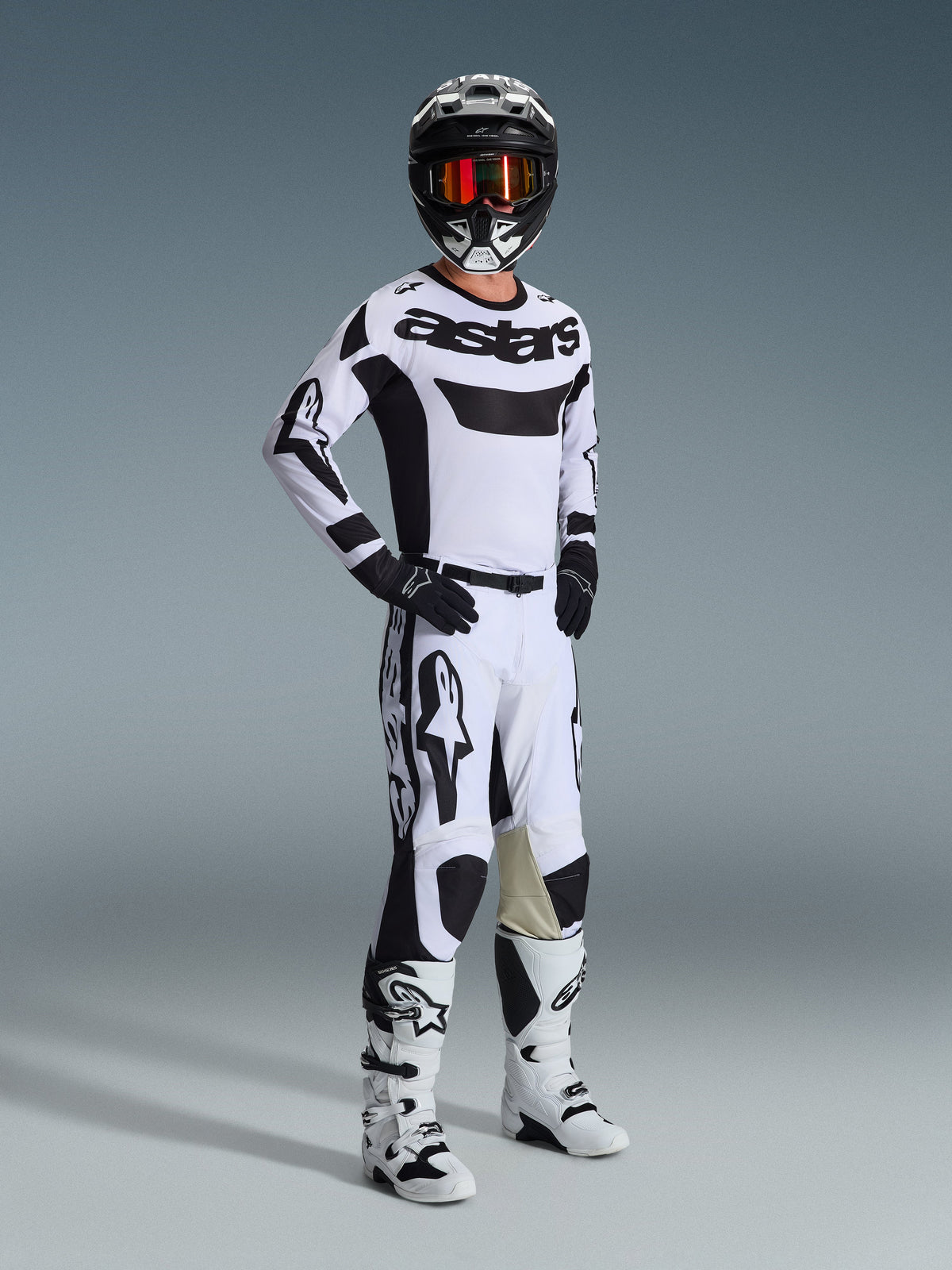 Motorcross rijder in complete race-uitrusting, Alpinestars 2026 Racer Riway Jersey in wit en zwart, bijpassende wit-zwarte motorcross broek, zwarte helm met gespiegelde bril, wit-zwarte laarzen en zwarte handschoenen, gecoördineerde outfit voor off-road racing
