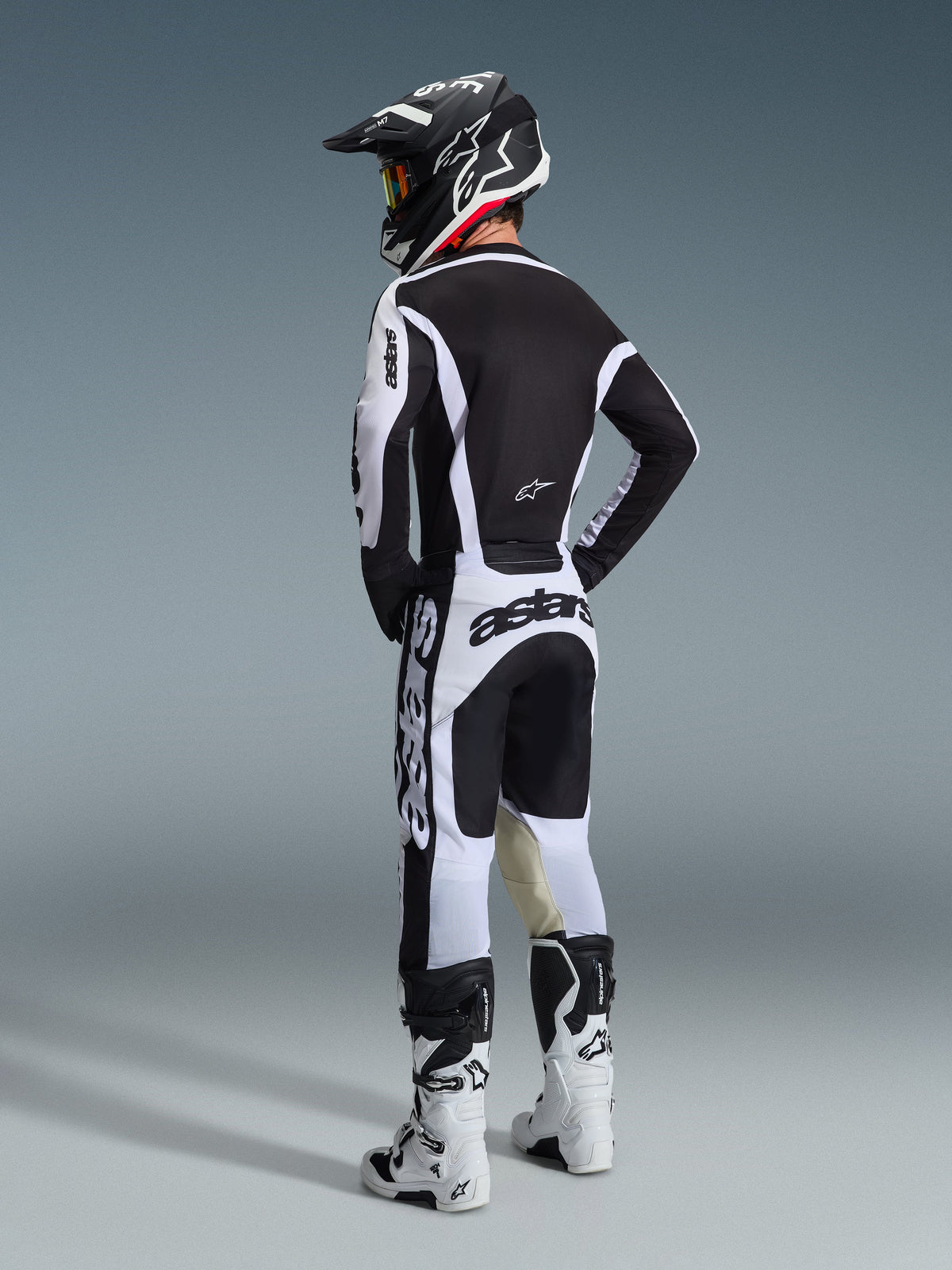 Motorcrossrijder in volledige race-uitrusting van achteren gezien, wit en zwart Alpinestars 2026 Racer Riway Jersey, bijpassende wit-zwarte broek met opvallende 'astars' branding, witte laarzen en een zwarte helm met witte accenten, gecoördineerde performance-outfit voor off-road racen.