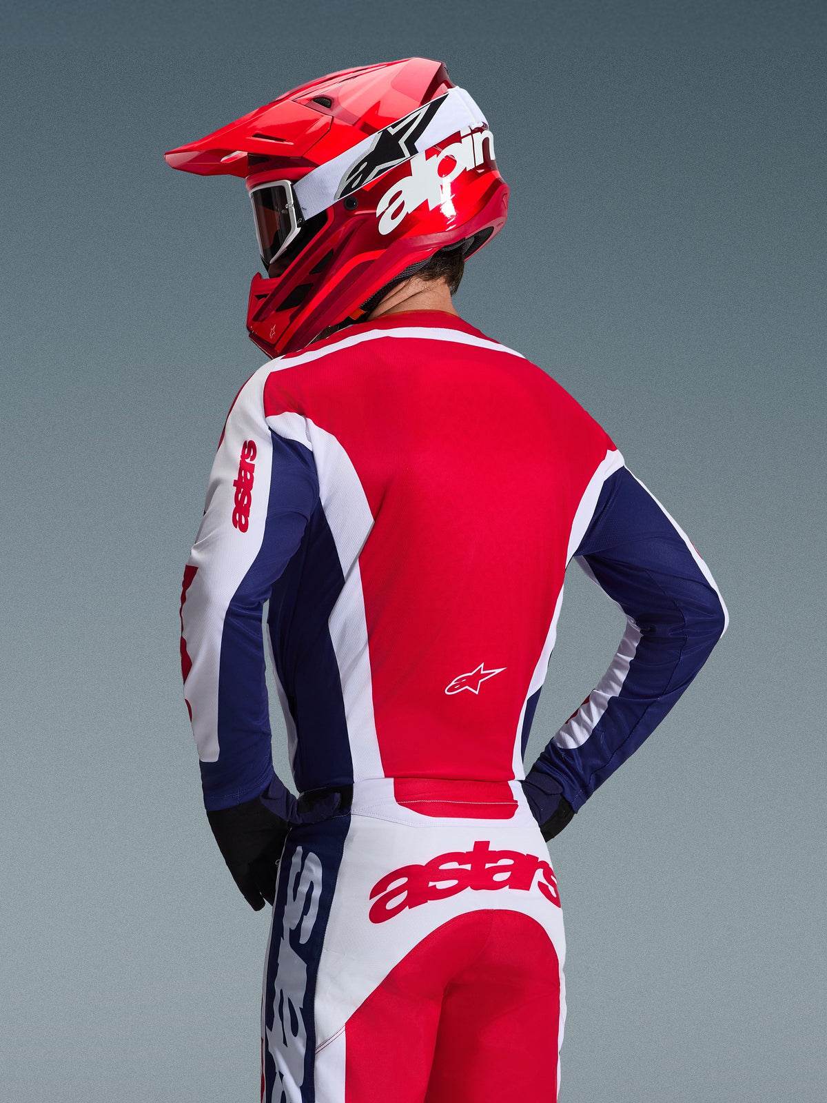 Motorcrossrijder van achteren in Alpinestars 2026 Racer Riway Jersey, kleurstelling wit, blauw en rood, bijpassende off-road broek met opvallende astars-branding, rode helm met wit Alpinestars-logo, zwarte handschoenen, gecoördineerde motorcross race-uitrusting