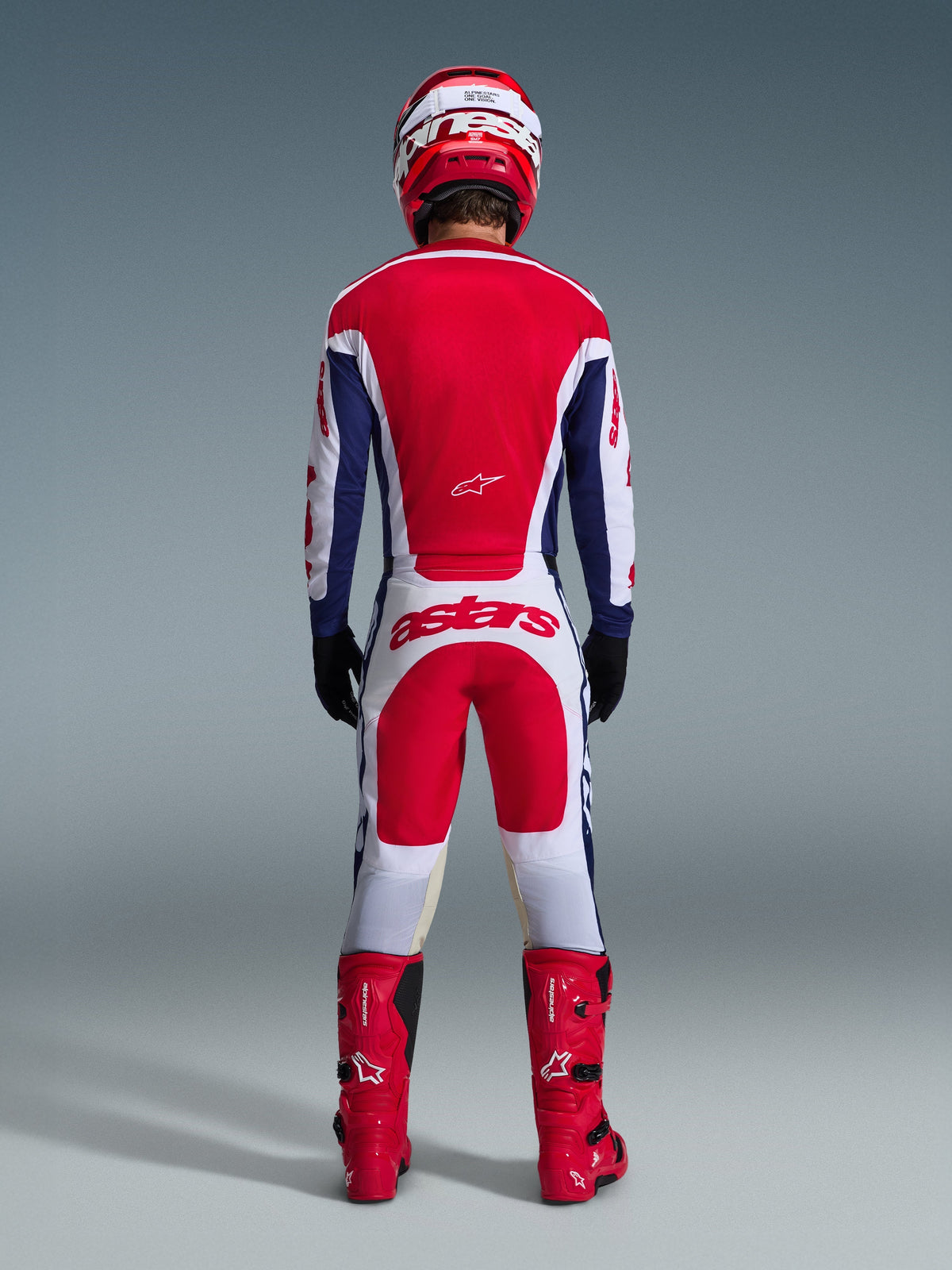 Motorcrossrijder in volledige race-uitrusting van achteren gezien, Alpinestars 2026 Racer Riway Jersey in White Blue Red, bijpassende witte en rode broek met opvallende 'astars' branding, rode laarzen en een rood-witte helm, gecoördineerde off-road race-outfit.