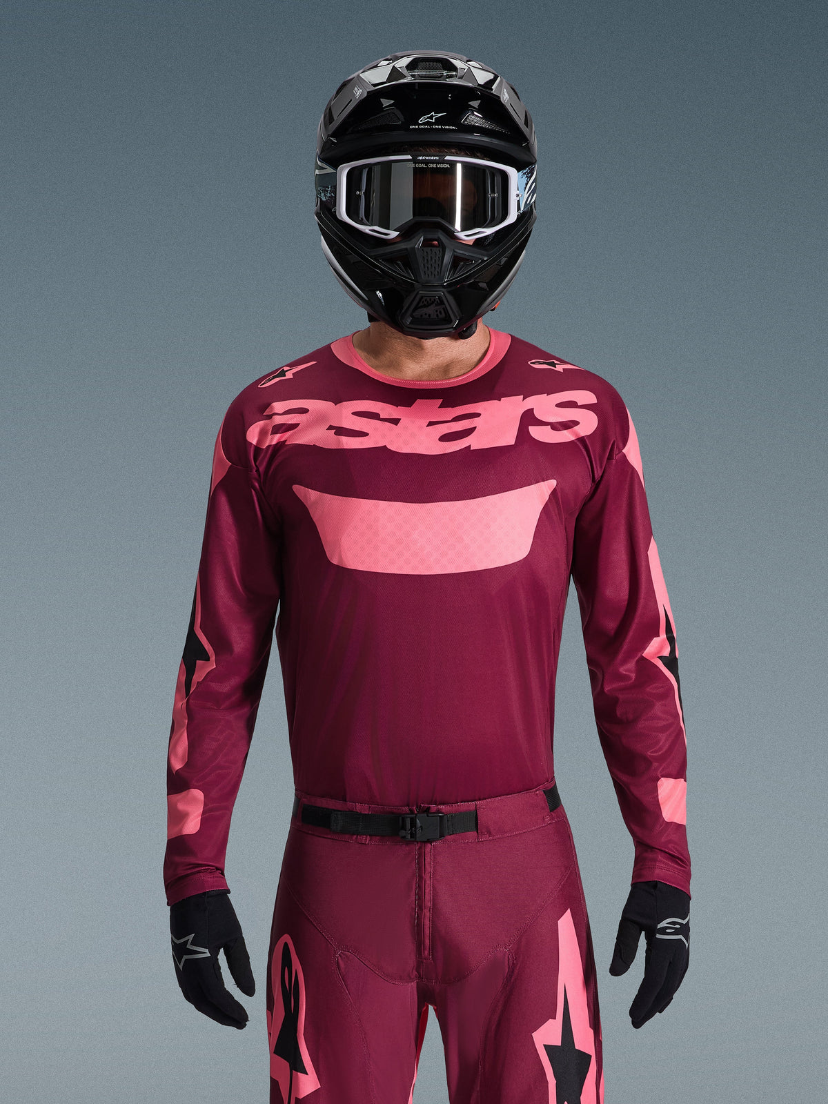 Motocross rider wearing Alpinestars 2026 Racer Riway Gear Set, Maroon and Pink, jersey with bold pink branding on the chest, matching pants with black and pink accents, black full-face helmet with goggles, and black gloves, coordinated off-road racing outfit.