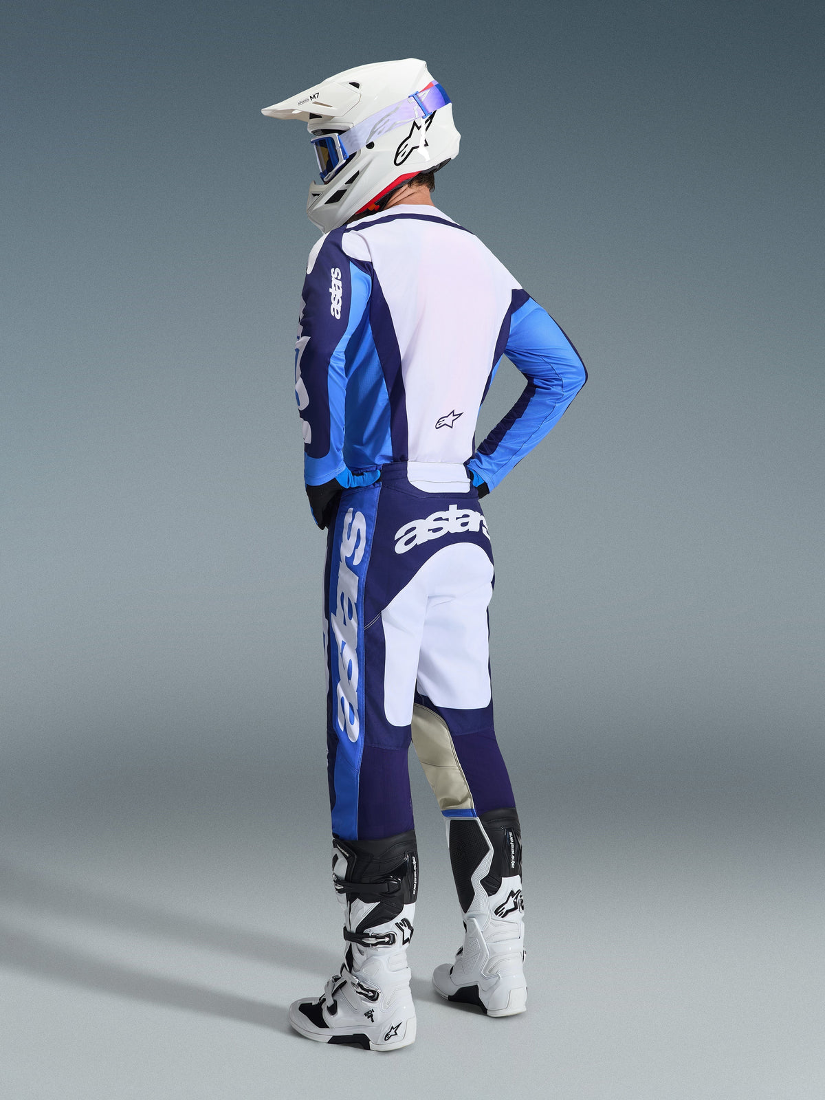 Motorcrosser van achteren gezien in de Alpinestars 2026 Racer Riway Gear Set in UCLA Blue en wit, bestaande uit een blauw-wit jersey met 'astars' branding, bijpassende blauw-witte broek, een witte motorcrosshelm met blauwe bril en wit-zwarte Tech-serie laarzen.