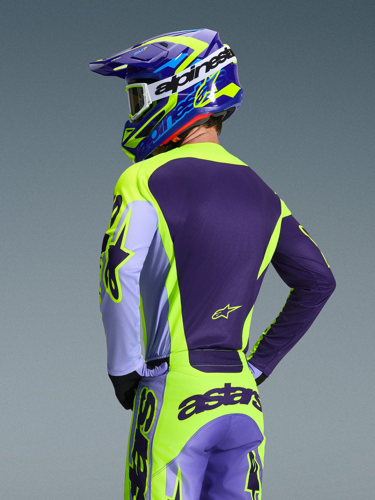 Alpinestars 2026 Racer Portl Jersey en bijpassende broek, motorcrosser van achteren gezien, paars en fluorgeel kleurenschema met lavendelkleurige accenten, bijpassende blauwe en gele helm, race-pasvorm uitrusting voor off-road prestaties