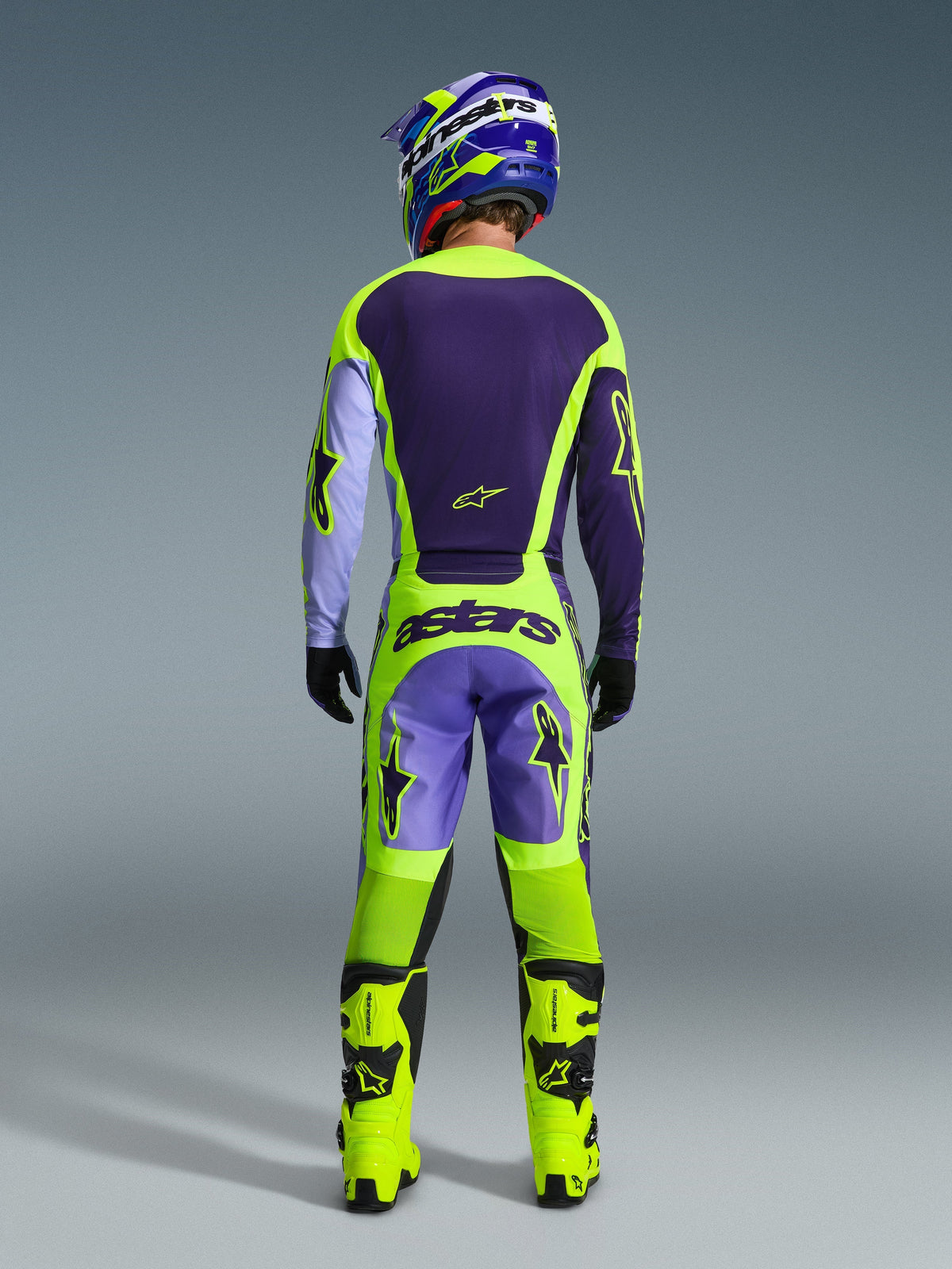 Motocrosser van achteren gezien in de Alpinestars 2026 Racer Portl Jersey in paars en fluo geel, gecombineerd met bijpassende motorcrossbroek, fluo gele laarzen en een blauw-gele helm, toont een complete off-road uitrusting.