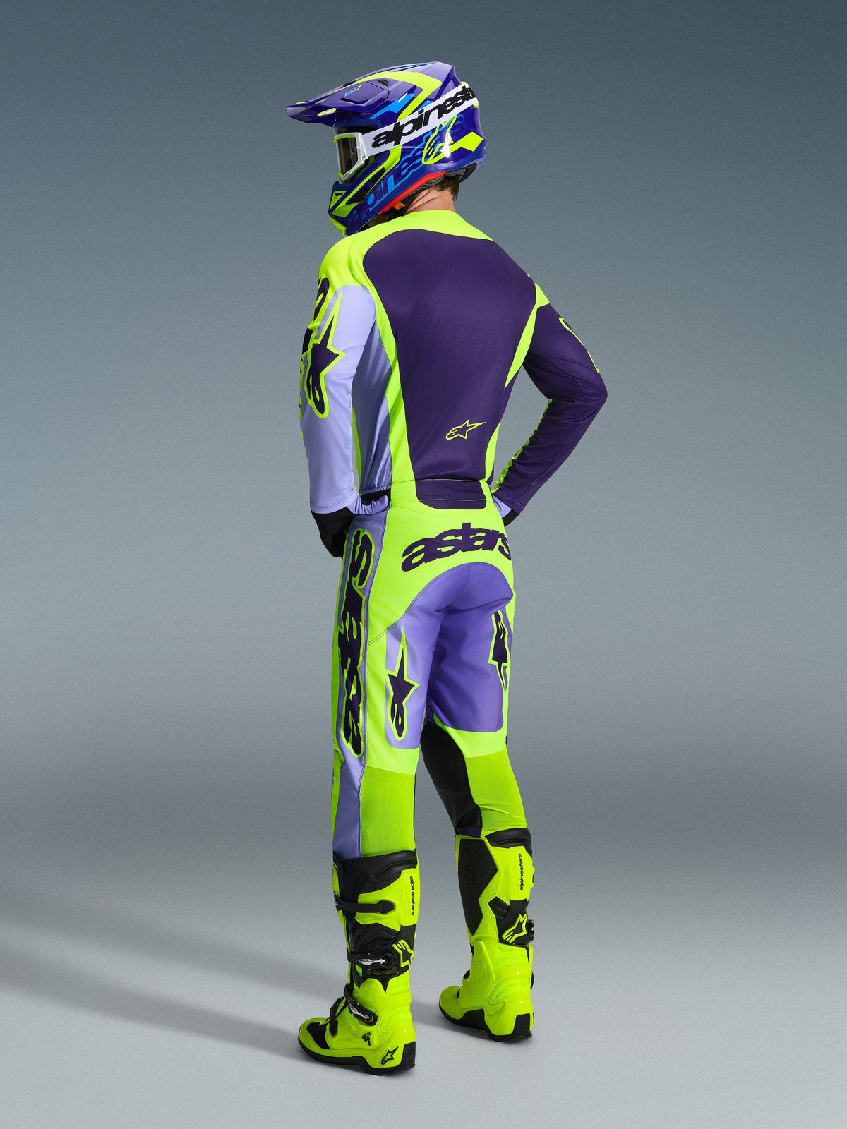 Motocrossrijder in Alpinestars 2026 Racer Portl Jersey en broek, paars en geel fluo kledingset, achteraanzicht met bijpassende helm en neongele laarzen, gecoördineerde professionele off-road race-outfit.