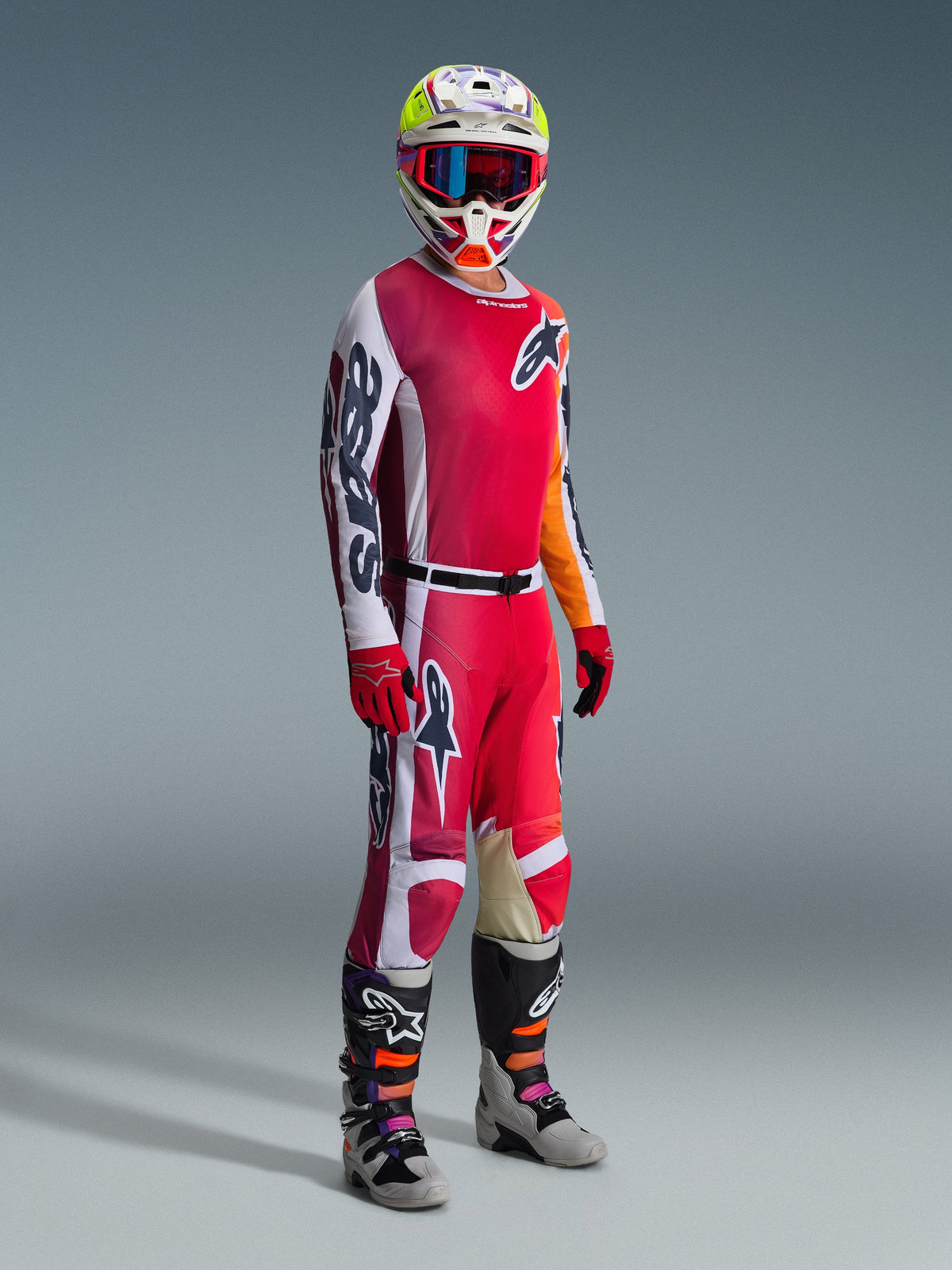 Motorcrosser in volledige race-uitrusting, Alpinestars 2026 Racer Portl Jersey in rood en lichtgrijs, bijpassende motorcrossbroek met witte en donkergroene accenten, meerkleurige helm met rode crossbril, grijze en zwarte laarzen met oranje en paarse details, rode handschoenen, gecoördineerde outfit voor off-road racen