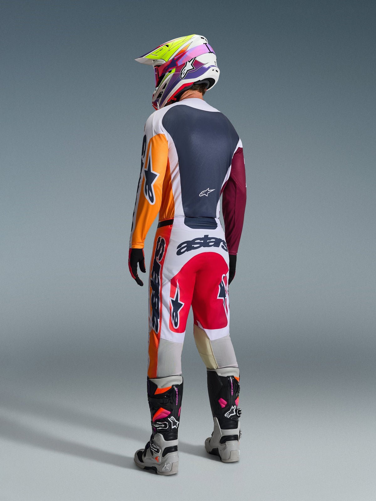 Motorcrosser in volledige race-uitrusting van achteren gezien, Alpinestars 2026 Racer Portl Jersey in Red Light Gray en Dark Green met oranje en kastanjebruine panelen, bijpassende witte en rode broek, grijze en zwarte laarzen met roze details, meerkleurige helm, gecoördineerde off-road race-outfit.